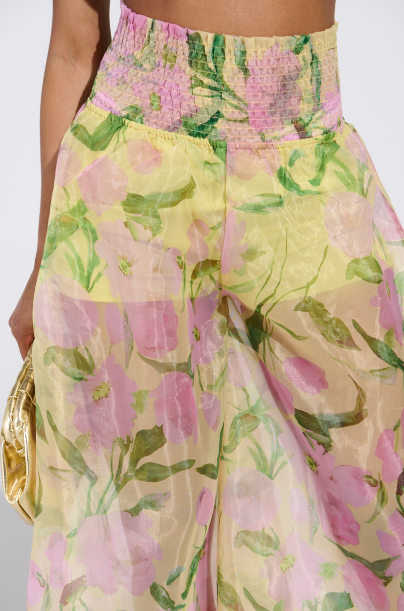 A person wearing the FLOWER POWER WIDE LEG PALAZZO PANT, featuring flowy chiffon fabric and a pastel floral pattern in pink and yellow, holds a gold clutch. The vibrant shirred waistband stands out against a soft white background.