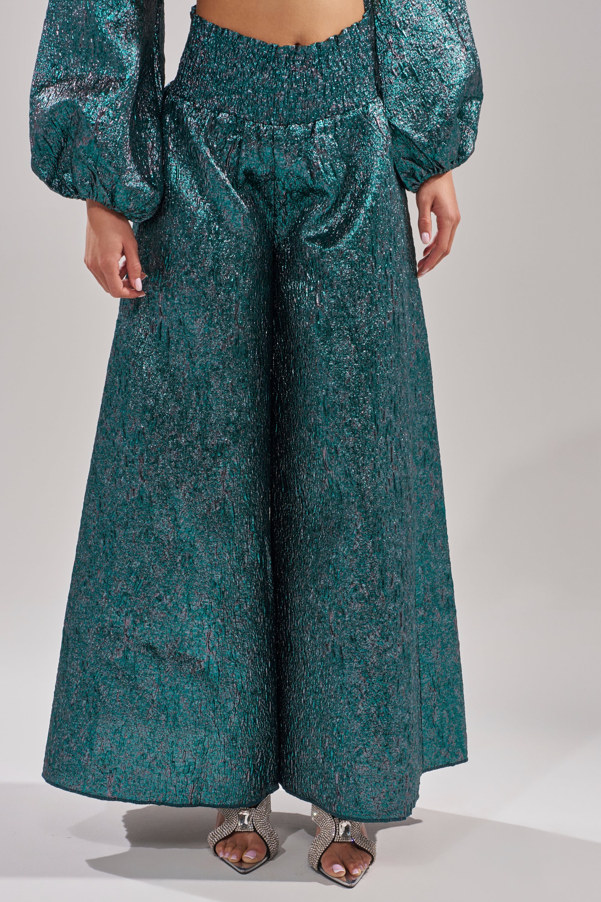 A person wears the ITS TIME TO CELEBRATE PALAZZO PANT in shiny, textured teal, paired with a matching puffy-sleeve top and silver open-toe heels. The image is cropped at the waist and ankles. 1