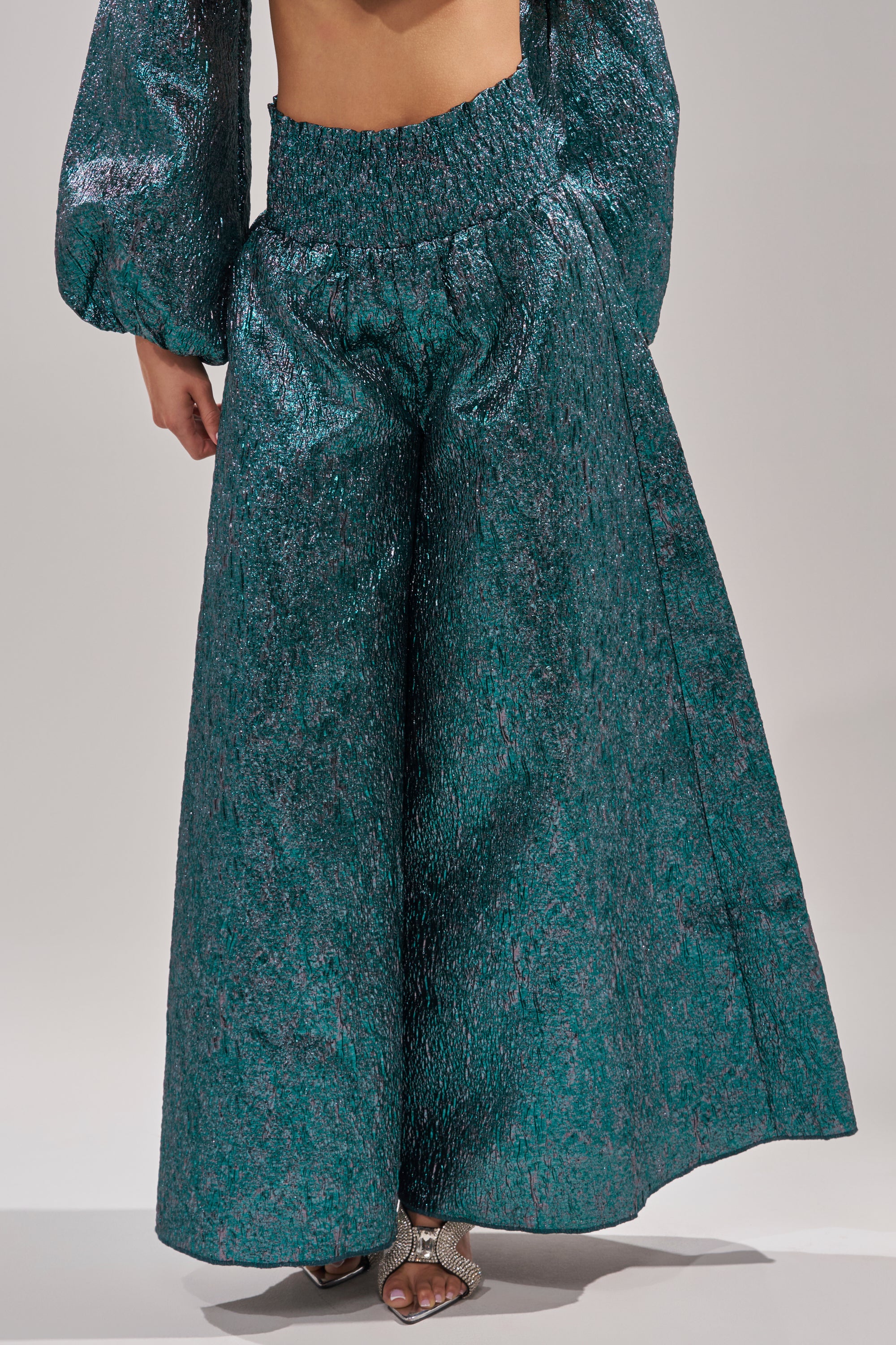 A person wears the ITS TIME TO CELEBRATE PALAZZO PANT in teal brocade with a metallic sheen, styled with a matching long-sleeve top and chic silver open-toe heels. 2