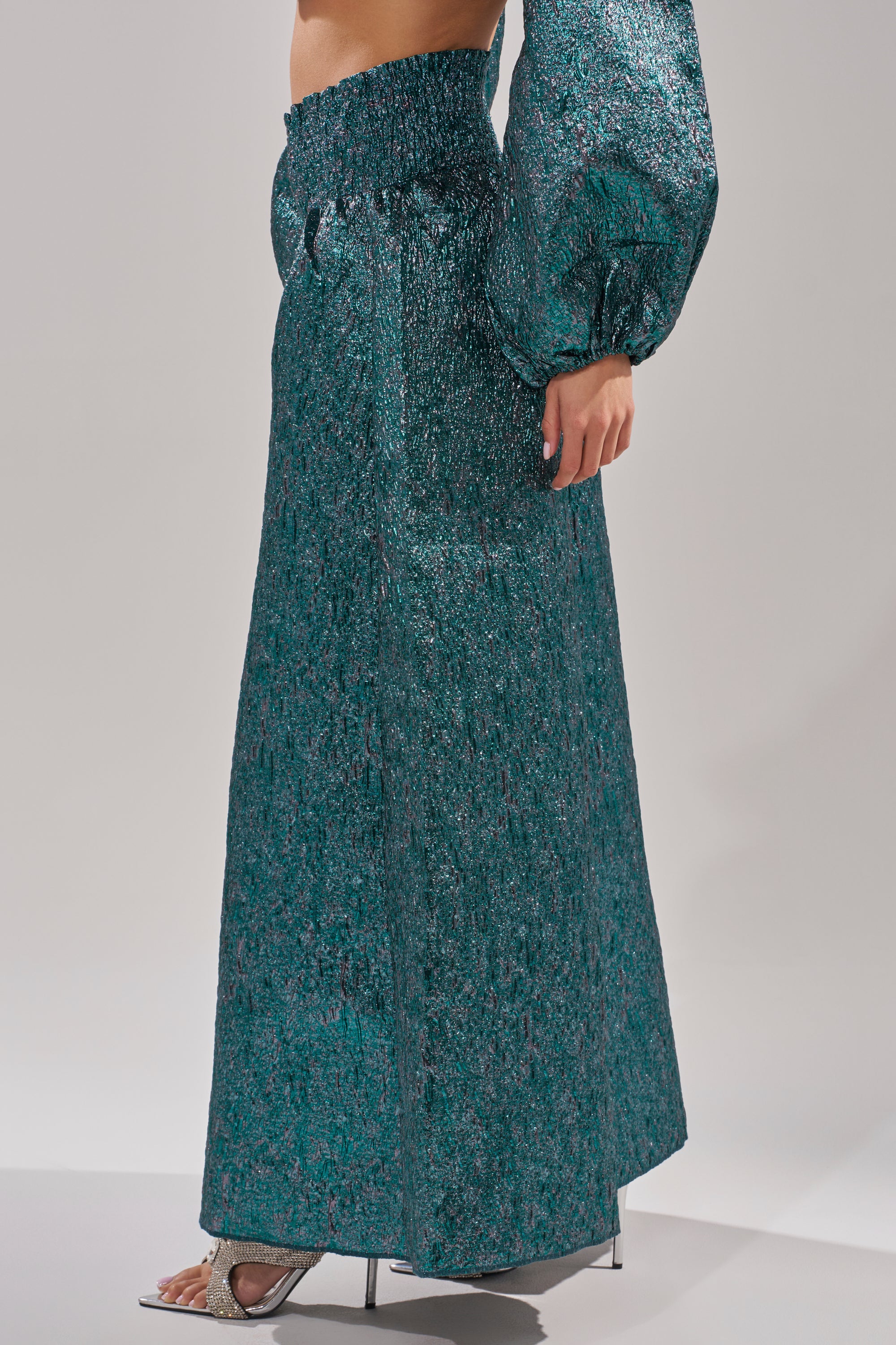 A person wears the ITS TIME TO CELEBRATE PALAZZO PANT in shimmering teal brocade with a ruffled high-rise waist, paired with a matching long-sleeve top and silver open-toe heels, standing against a plain light gray background. 3