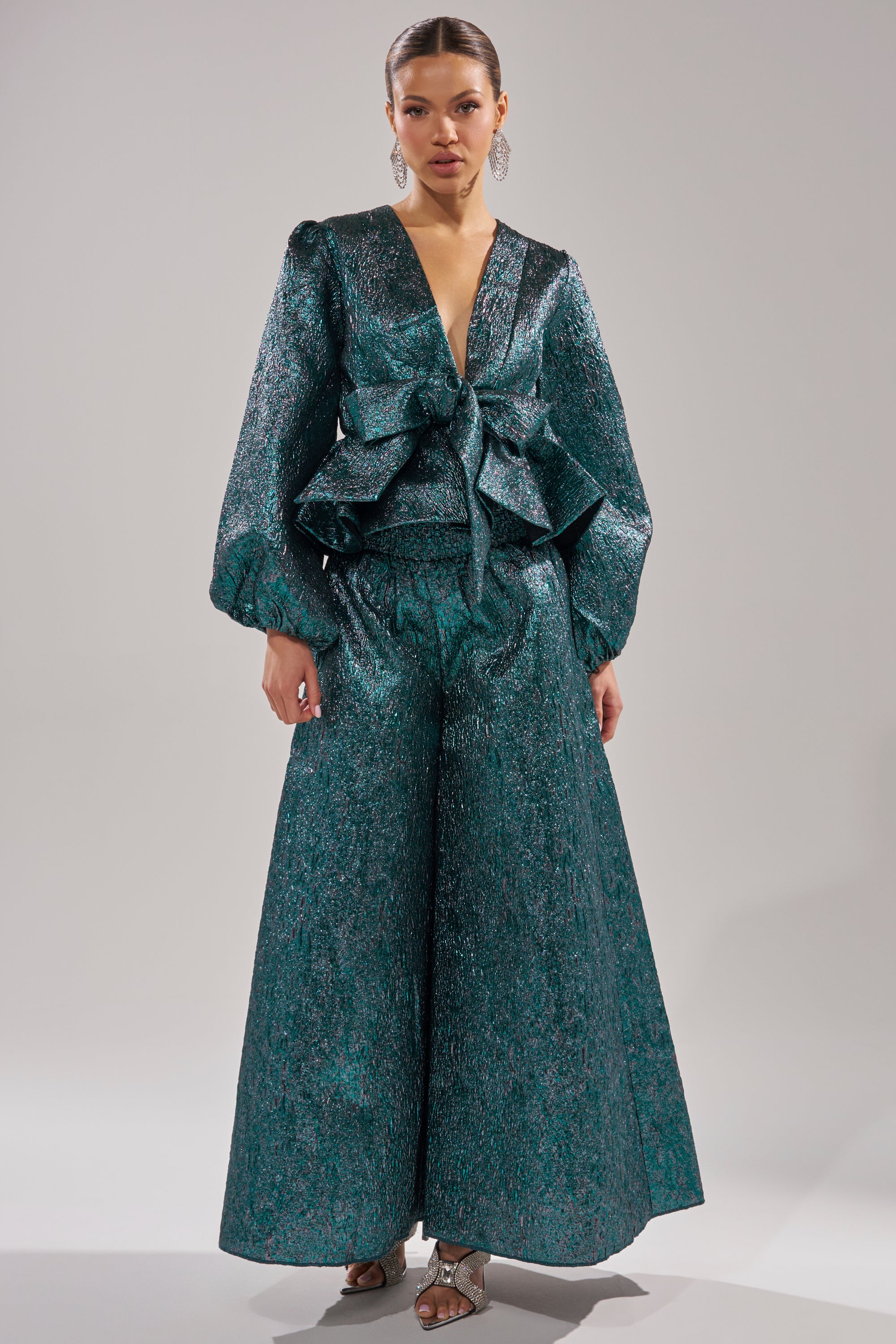A woman wears the ITS TIME TO CELEBRATE PALAZZO PANT, featuring shimmering teal brocade, high-rise palazzo legs, a deep V-neck top with a large waist bow, styled with dangling earrings and metallic heeled sandals. 5
