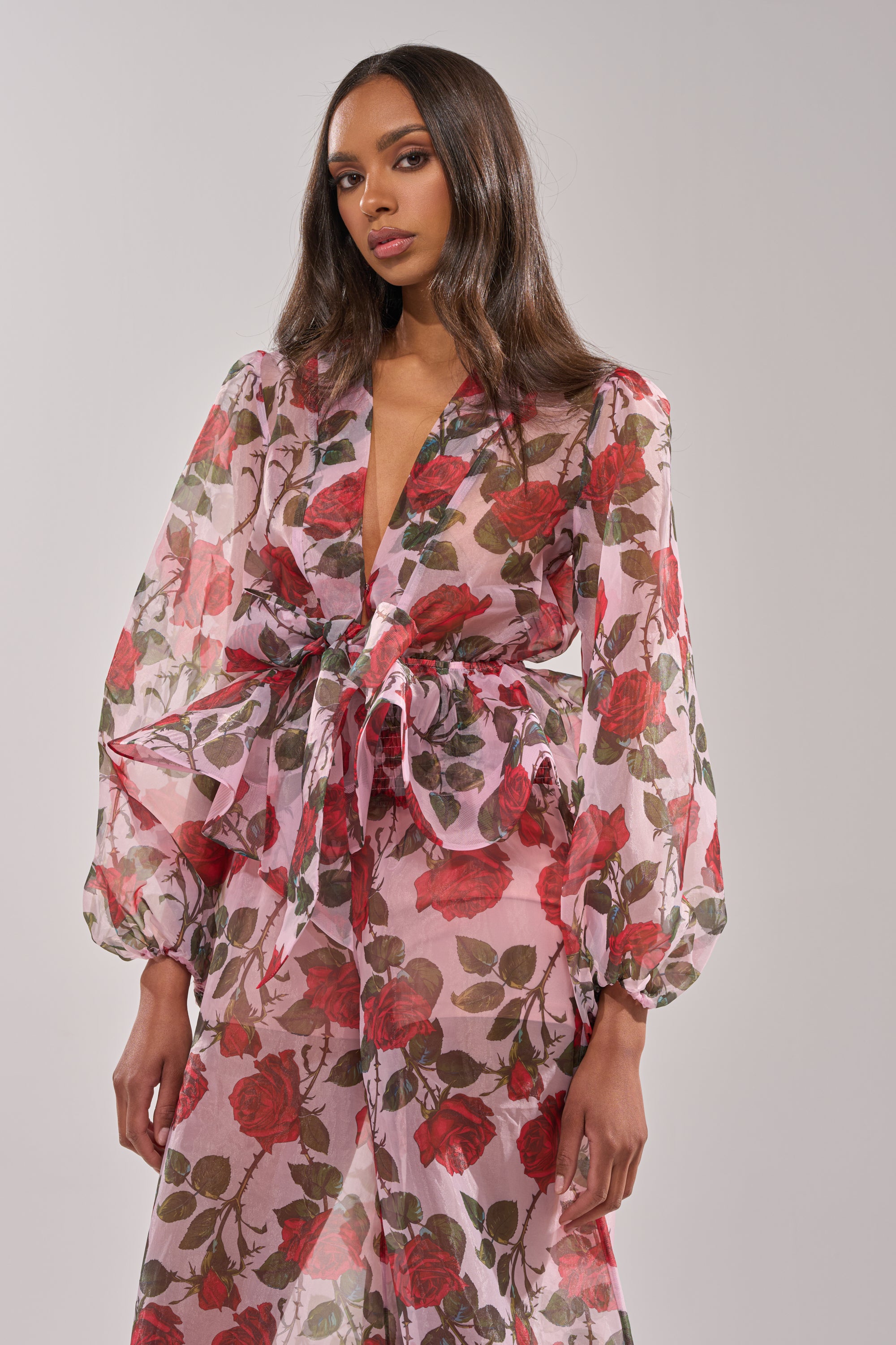 A woman models the PRETTY GIRL FLORAL BLOUSE—a sheer, long-sleeve top with a pink base, red rose print, deep V-neckline, and peplum silhouette—against a plain light background. 0