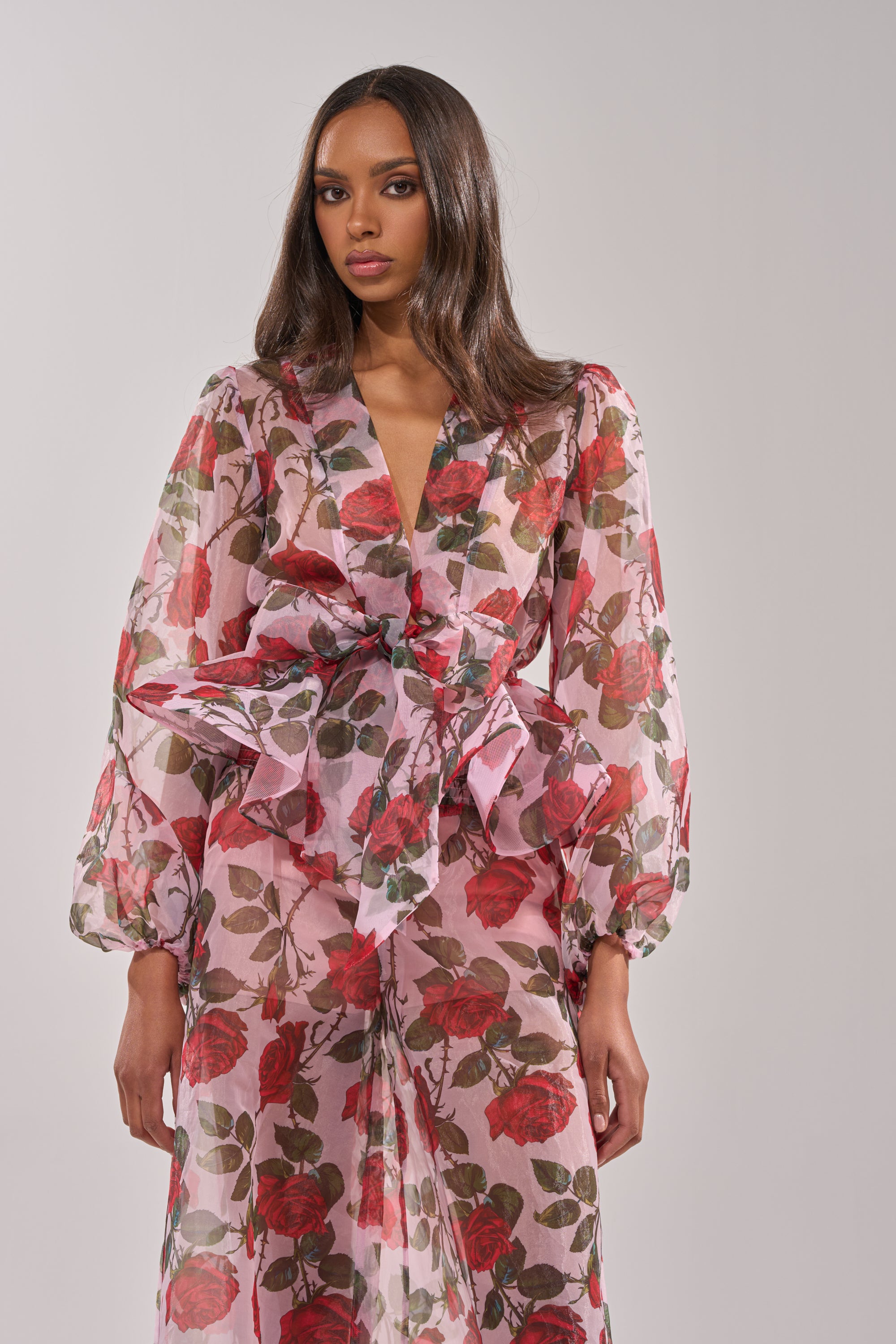 A woman models the PRETTY GIRL FLORAL BLOUSE—a sheer, long-sleeved pink top with red rose prints and a statement bow at the waist. Her long, straight hair stands out against a plain light background. 1
