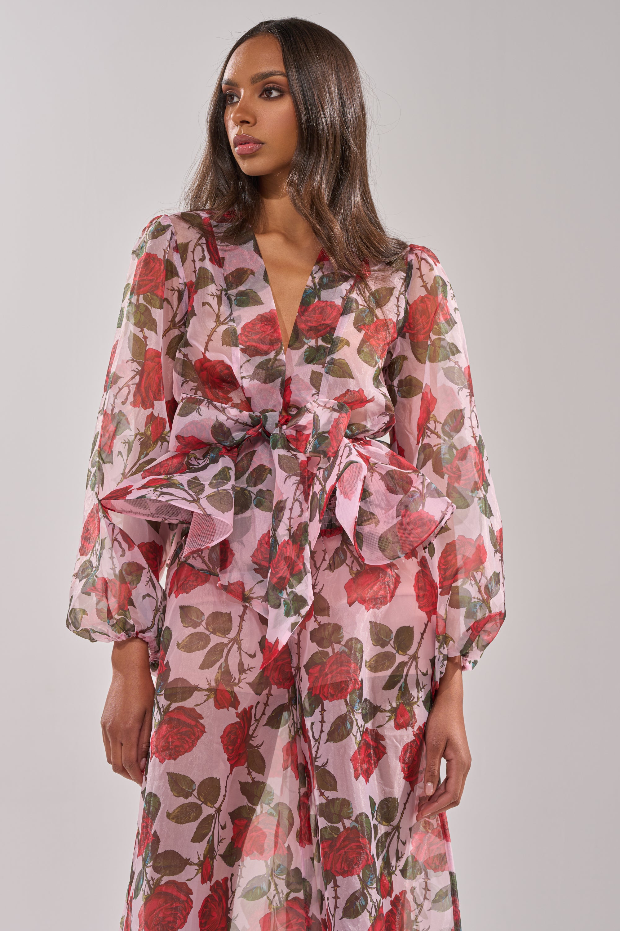A woman stands against a plain light gray background wearing the PRETTY GIRL FLORAL BLOUSE, featuring voluminous sleeves and a large waist bow, paired with a long, sheer pink skirt. 2