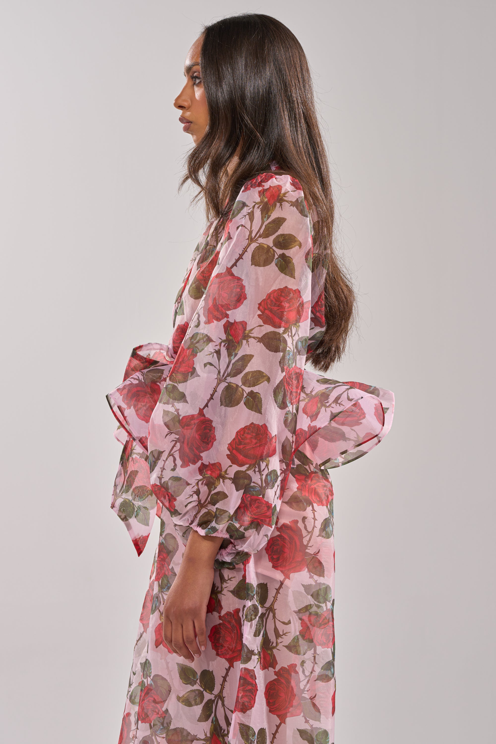 A woman with long dark hair is shown in profile against a plain light background wearing the PRETTY GIRL FLORAL BLOUSE, a sheer pink top featuring red rose and green leaf prints, voluminous sleeves, and a large waist bow. 3