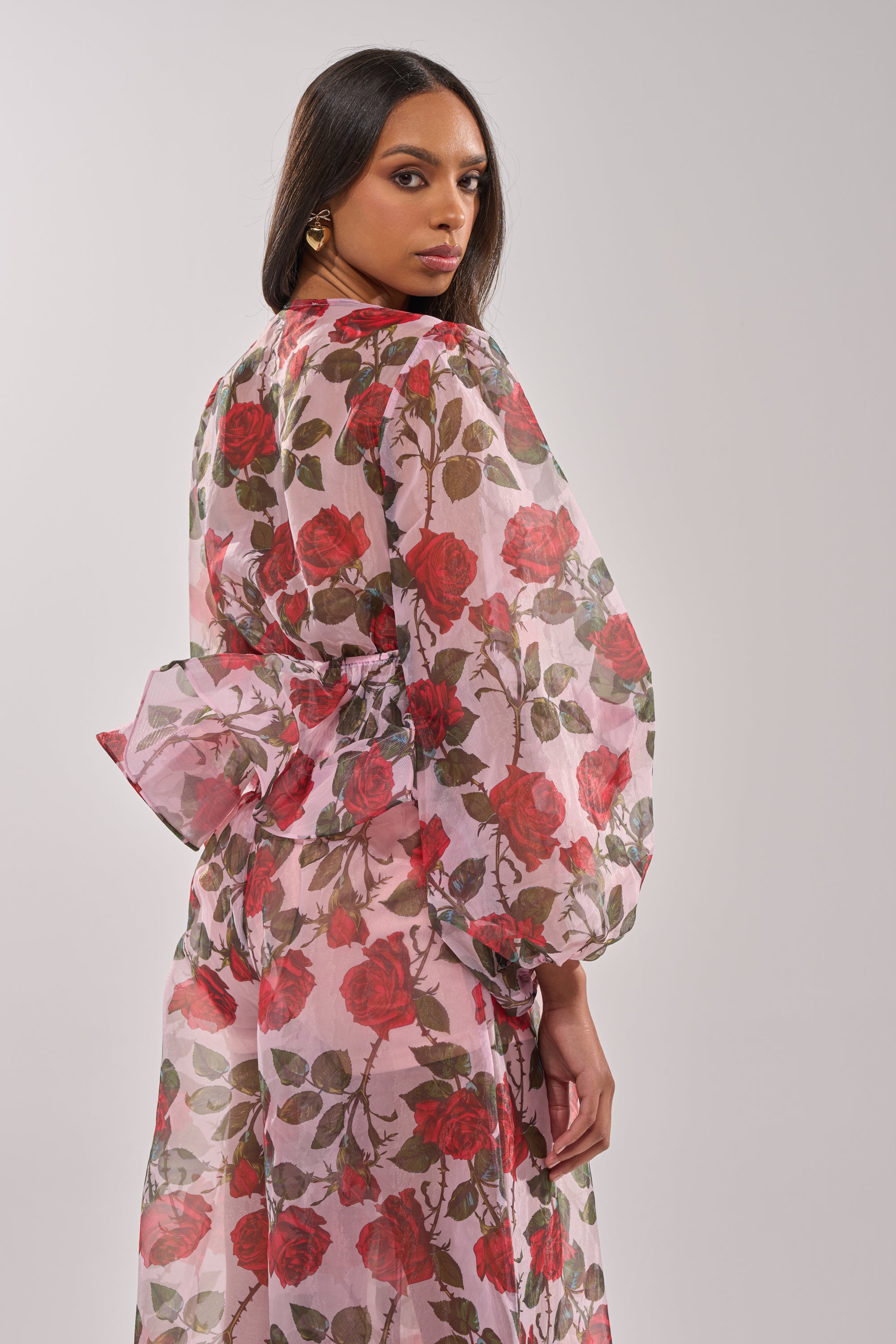 A woman in the PRETTY GIRL FLORAL BLOUSE—a sheer, long-sleeved rose print top with a large back bow—stands facing away, looking over her shoulder with a neutral expression against a plain background. 4