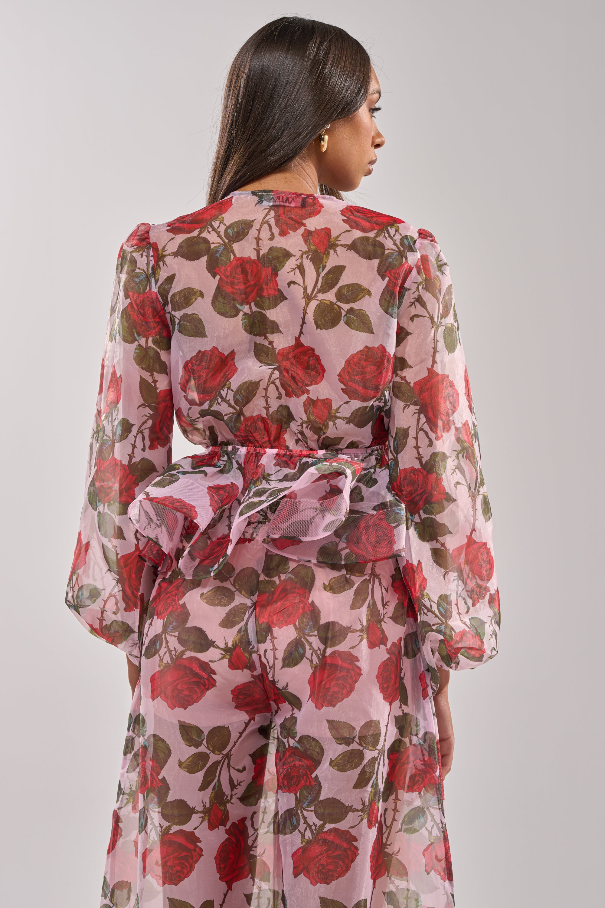 A woman faces away, wearing the PRETTY GIRL FLORAL BLOUSE—a sheer, long-sleeved dress with a large back bow. Its flowy pink base is adorned with a vivid red and green floral pattern featuring bold red roses. 5
