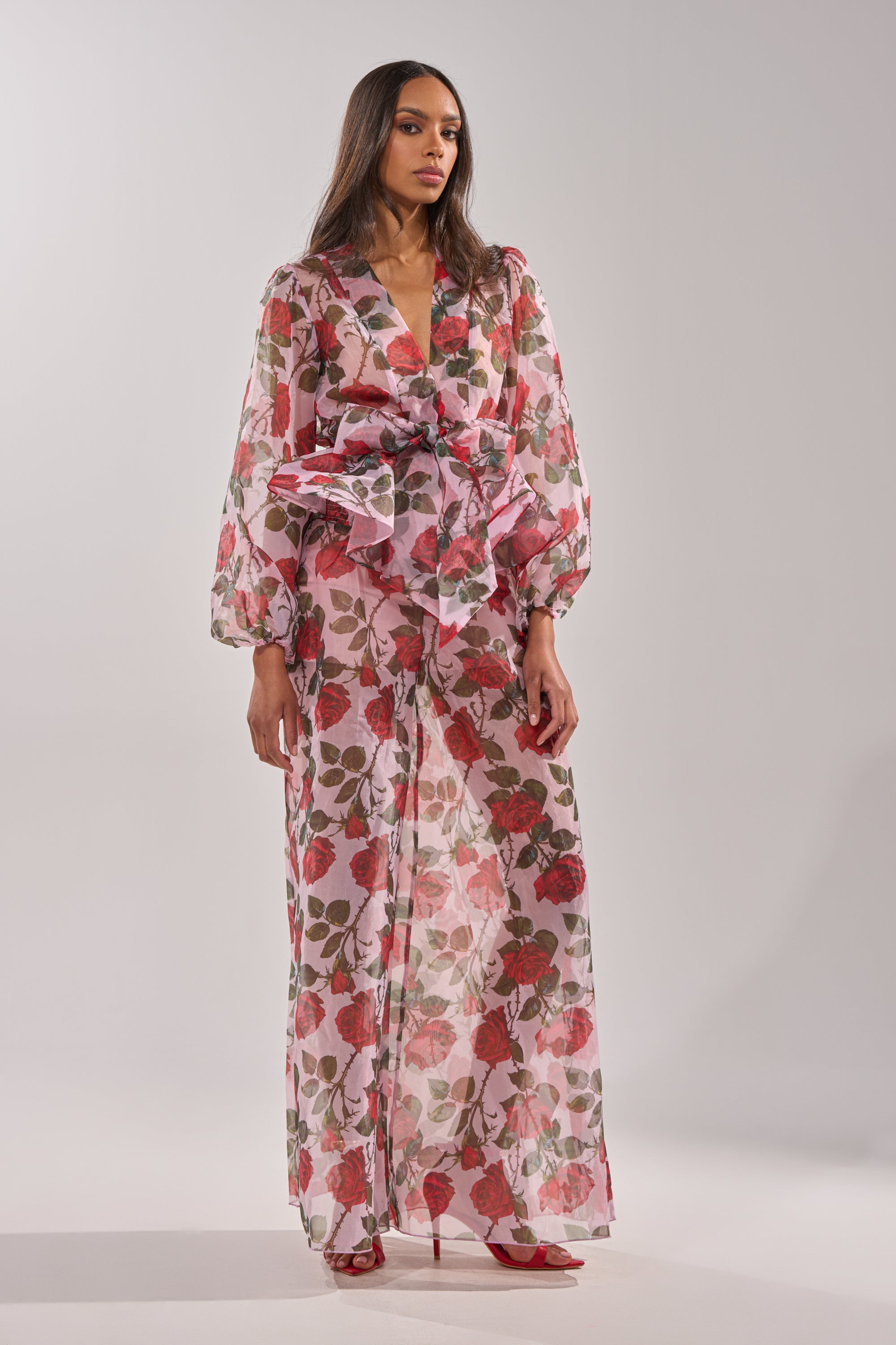 A woman wears the PRETTY GIRL FLORAL BLOUSE, a long, sheer rose print dress with puffed sleeves and a large bow at the waist. She has long hair and red heels, standing against a plain background. 7