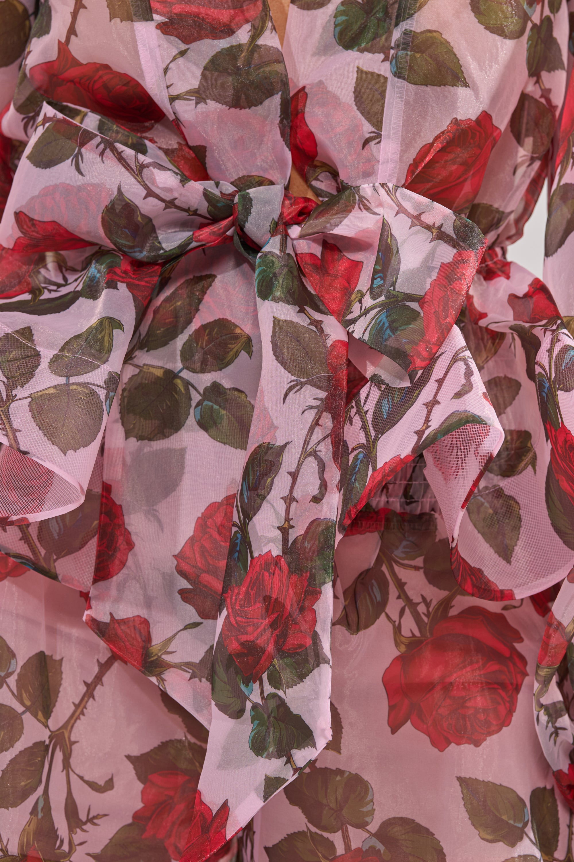 The PRETTY GIRL FLORAL BLOUSE is a sheer wrap top with red roses and green leaves on a light pink, slightly translucent fabric, finished with a large decorative bow for delicate charm. 8