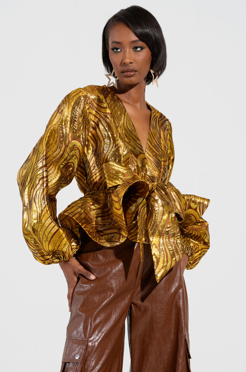 The individual stands confidently wearing the IN A DAYDREAM TIE FRONT BLOUSE IN GOLD, featuring a large bow and wide sleeves, coupled with metallic knit brown leather pants. They accessorize with star-shaped earrings and maintain a neutral expression against a plain background.