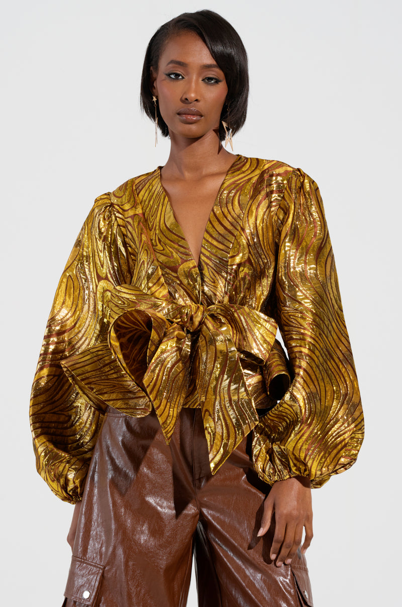 A person confidently stands against a white background, wearing an "IN A DAYDREAM TIE FRONT BLOUSE IN GOLD," paired with brown leather pants. Their hair is styled short, and they wear hoop earrings.