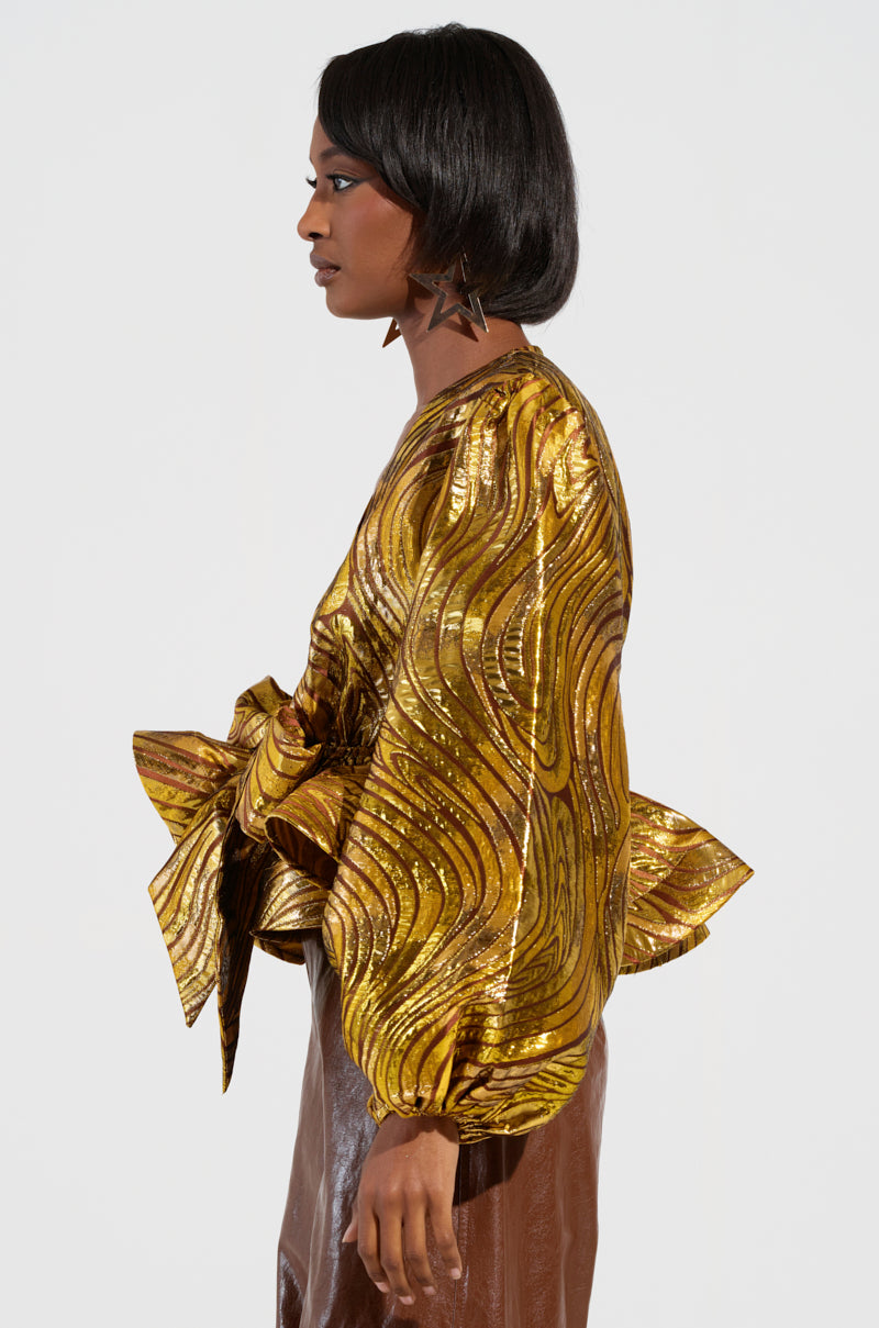 A person stands in profile wearing the "In a Daydream Tie Front Blouse in Gold," featuring a shimmering peplum silhouette, textured wavy pattern, and large bows on the sleeves. They have short black hair and star-shaped earrings, set against a plain white background.