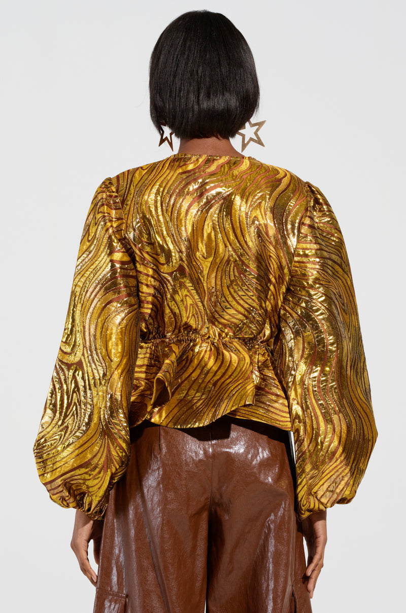 A person with short hair is facing away, wearing the IN A DAYDREAM TIE FRONT BLOUSE IN GOLD, known for its shimmering metallic knit fabric and swirling patterns, paired beautifully with their brown pants that have a shiny texture and adorned with star-shaped earrings.