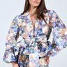 A woman with long, straight hair and hoop earrings poses in the EXALTED FLORAL TIE FRONT BLOUSE featuring a colorful floral pattern, V-neck, and waist bow. The peplum silhouette adds elegance against a plain backdrop.