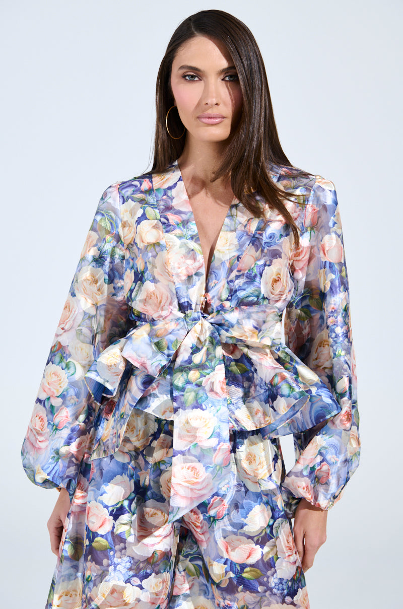 A woman showcases her style in the EXALTED FLORAL TIE FRONT BLOUSE with matching pants, featuring long sleeves and a waist tie for a peplum silhouette. She accessorizes with large hoop earrings against a plain light gray background.