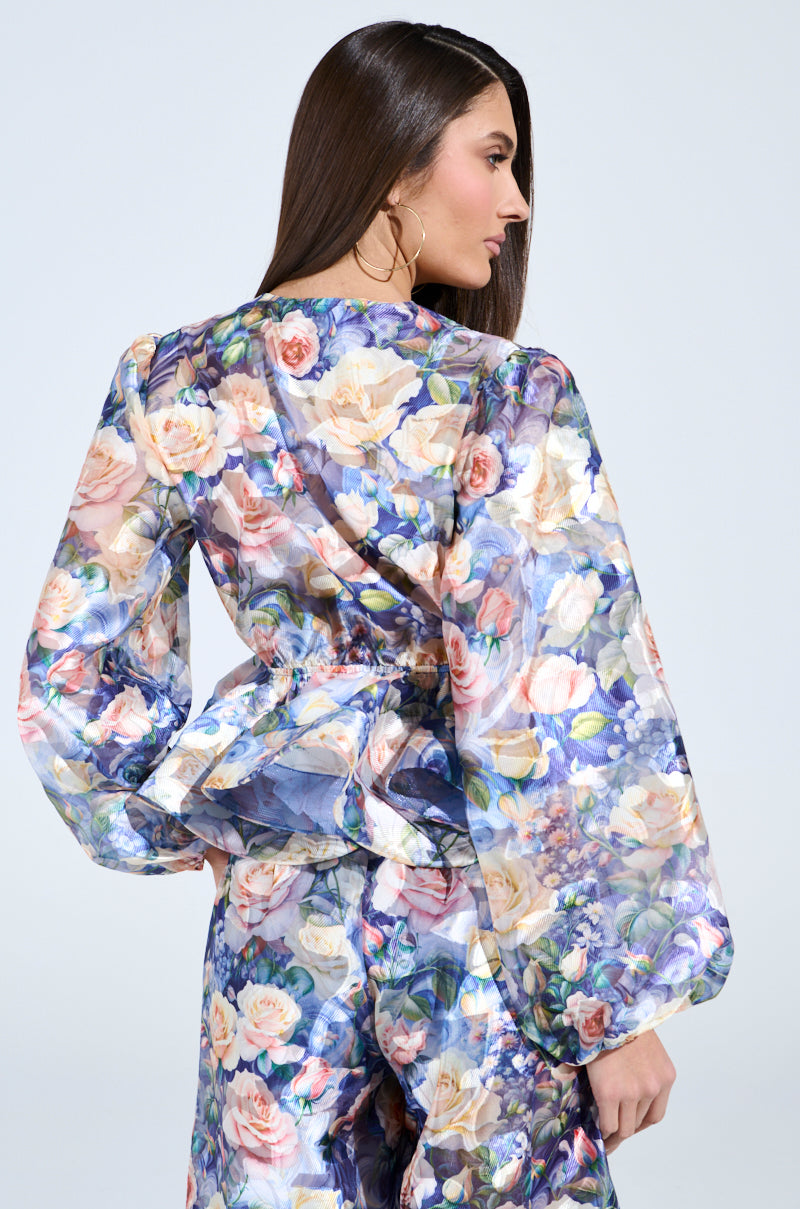 A woman with long brown hair wears the EXALTED FLORAL TIE FRONT BLOUSE, featuring sheer mesh fabric. The long sleeves and peplum silhouette add elegance, while a plain backdrop highlights the vibrant floral design.