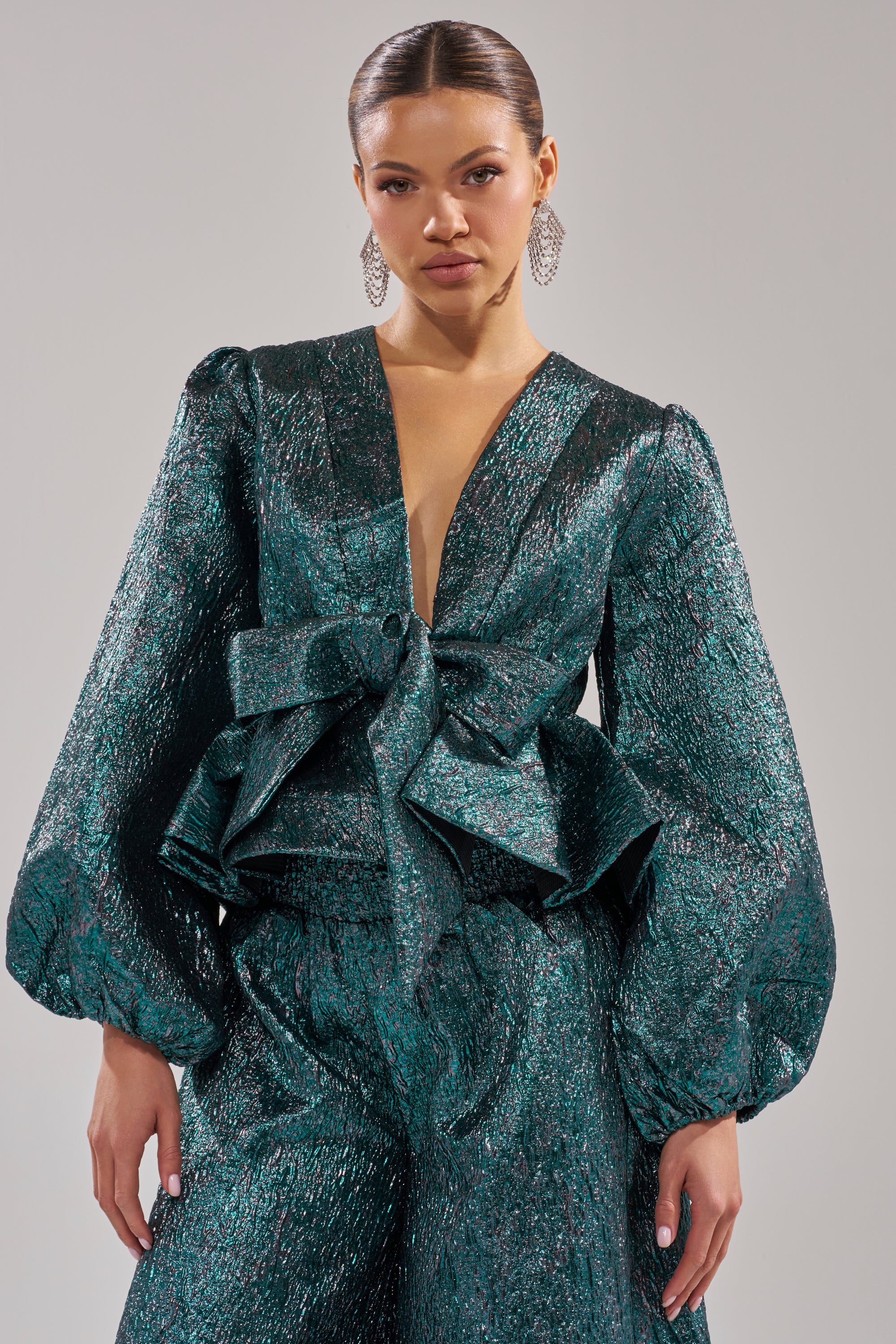 A woman poses against a plain light background wearing the jewel-toned ITS TIME TO CELEBRATE BLOUSE, styled with a textured deep green metallic pantsuit featuring voluminous sleeves, a plunging neckline, and a statement waist bow. 0