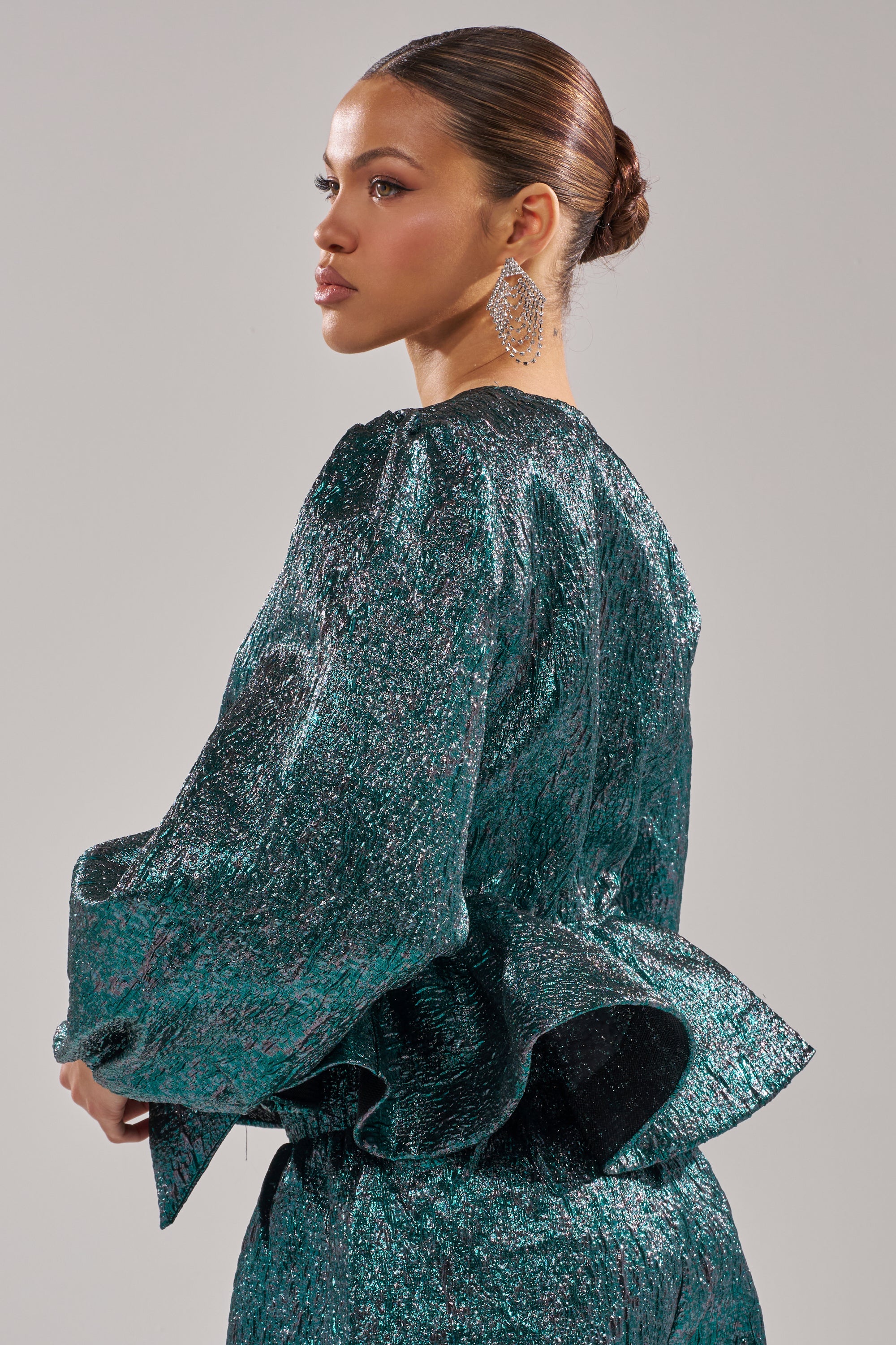 A woman with her hair in a sleek bun wears the ITS TIME TO CELEBRATE BLOUSE, a teal green brocade top with voluminous sleeves and a peplum silhouette. She’s shown in profile, highlighting statement drop earrings against a plain background. 3