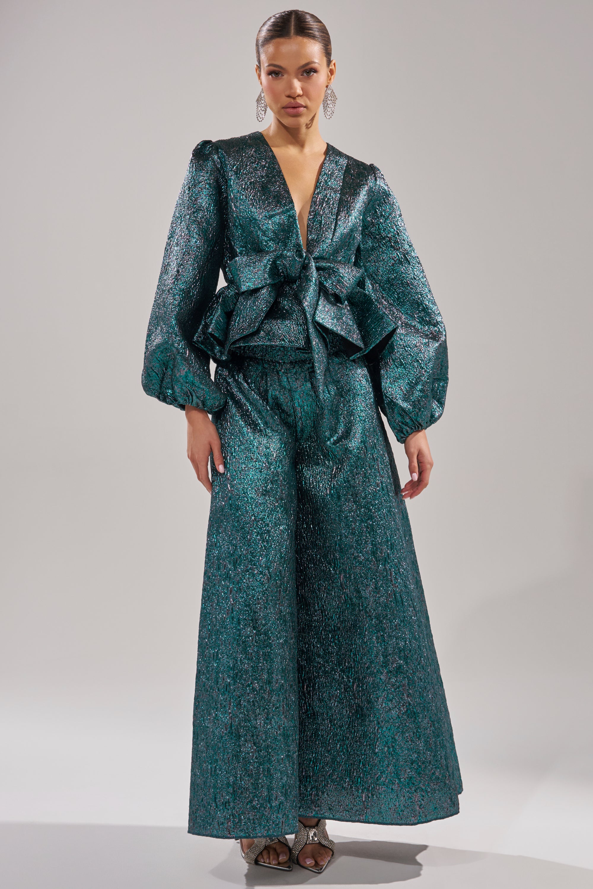 A woman wears the ITS TIME TO CELEBRATE BLOUSE in deep green brocade, featuring a plunging neckline, voluminous sleeves, and a large waist bow. She faces forward against a plain light background, paired with wide-leg pants. 5