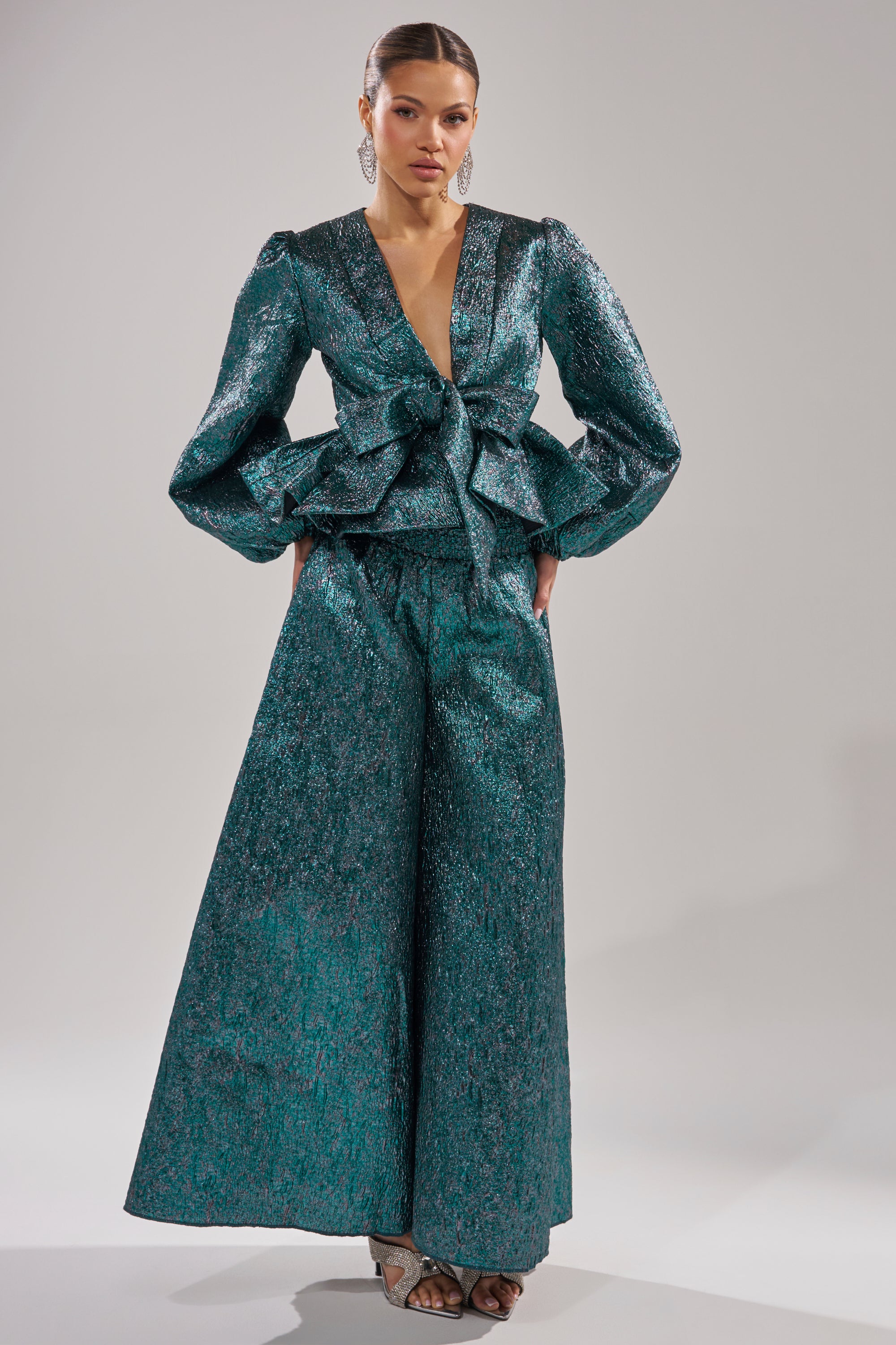 A woman wears the ITS TIME TO CELEBRATE BLOUSE—a shiny teal, deep V-neck, long-sleeve top with a large front bow—paired with wide-leg trousers and heels, posing against a plain background. 6