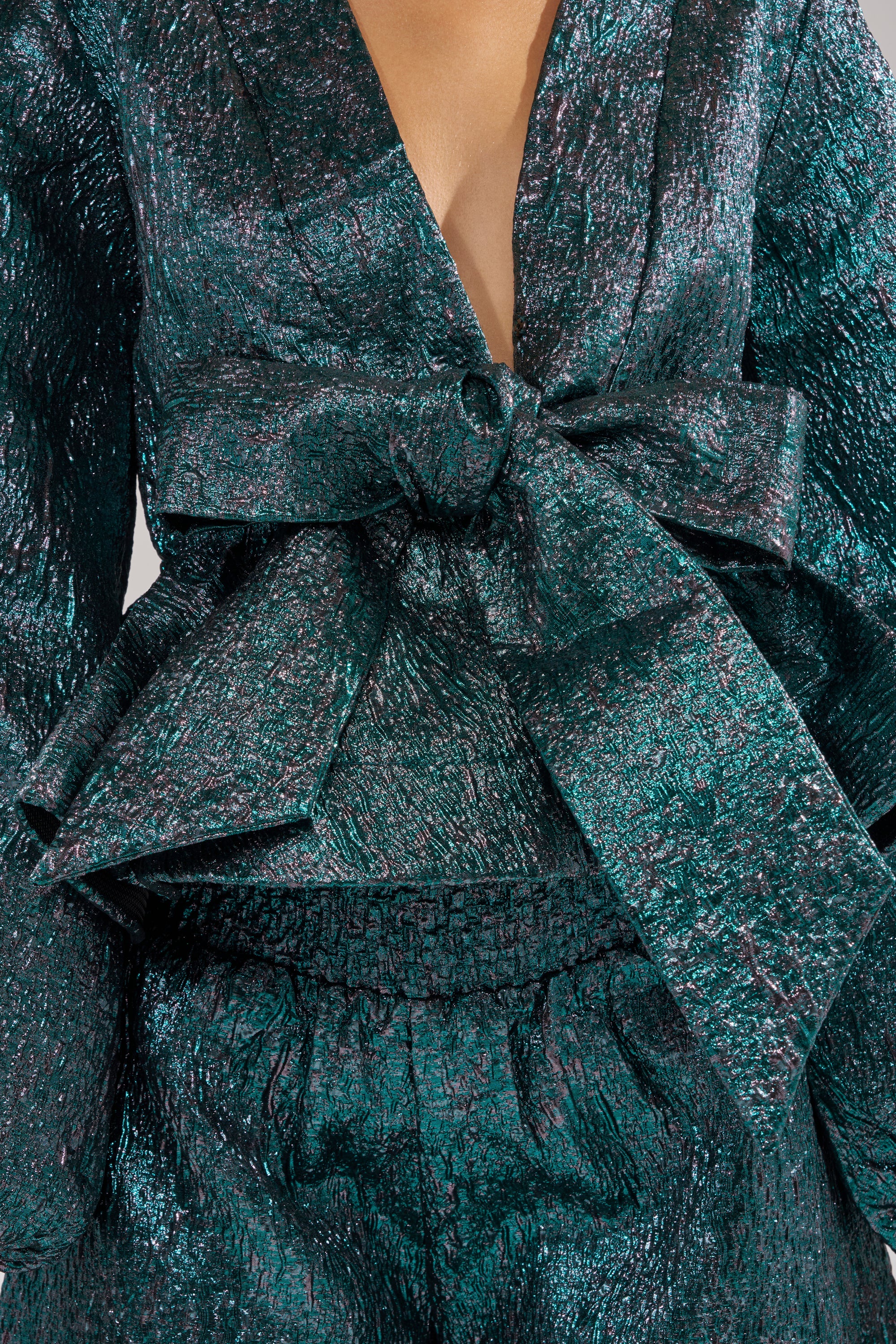 A close-up of a person wearing the ITS TIME TO CELEBRATE BLOUSE, a metallic teal brocade top with a deep V-neckline, crinkled shimmering fabric, and a large waist bow. The face is not visible. 7