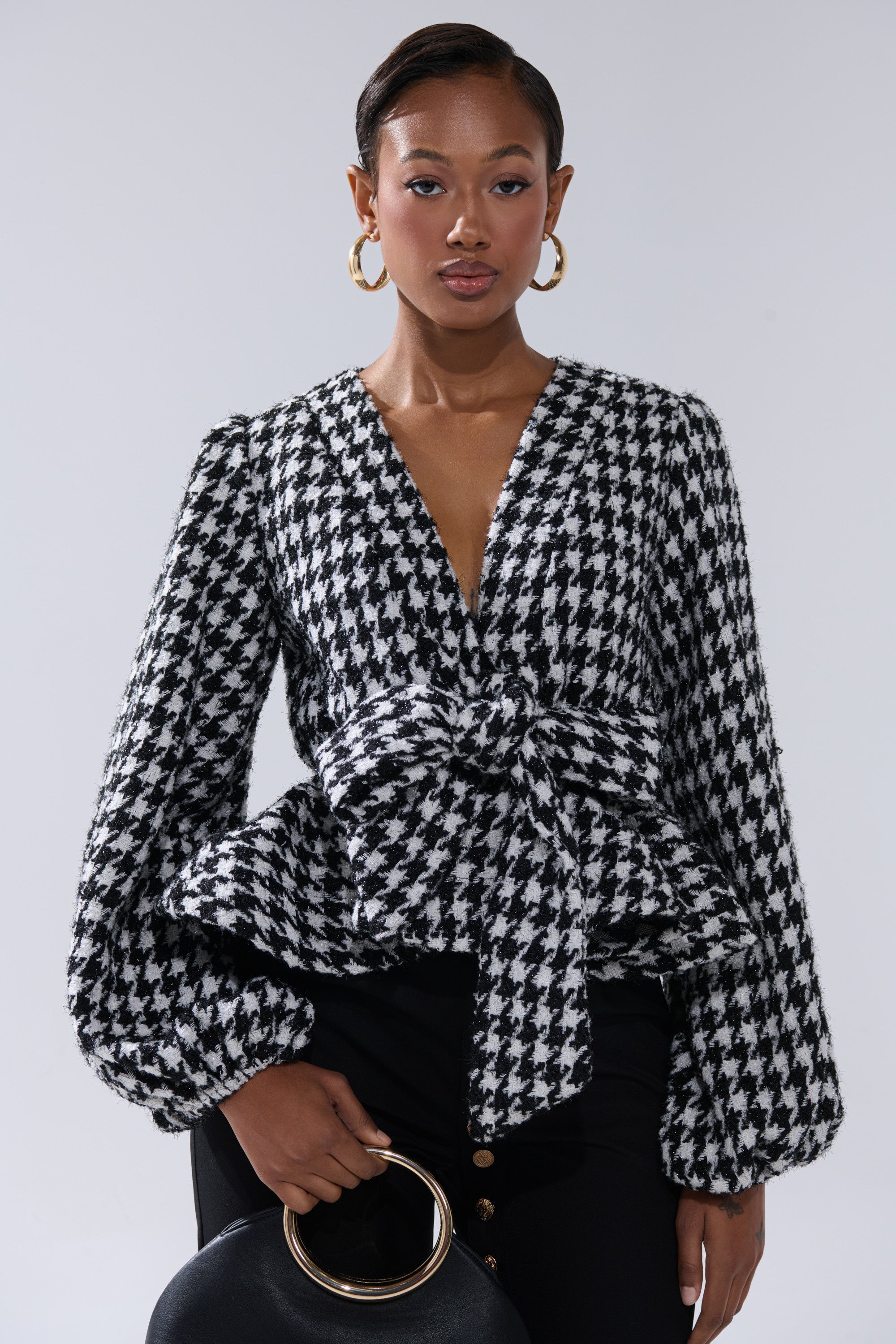 A woman wears the GIRL PLEASE HOUNDSTOOTH TIE-FRONT BLOUSE with balloon sleeves, peplum hem, and a statement waist bow, styled with black pants, gold hoop earrings, and a chic black handbag featuring a gold handle.