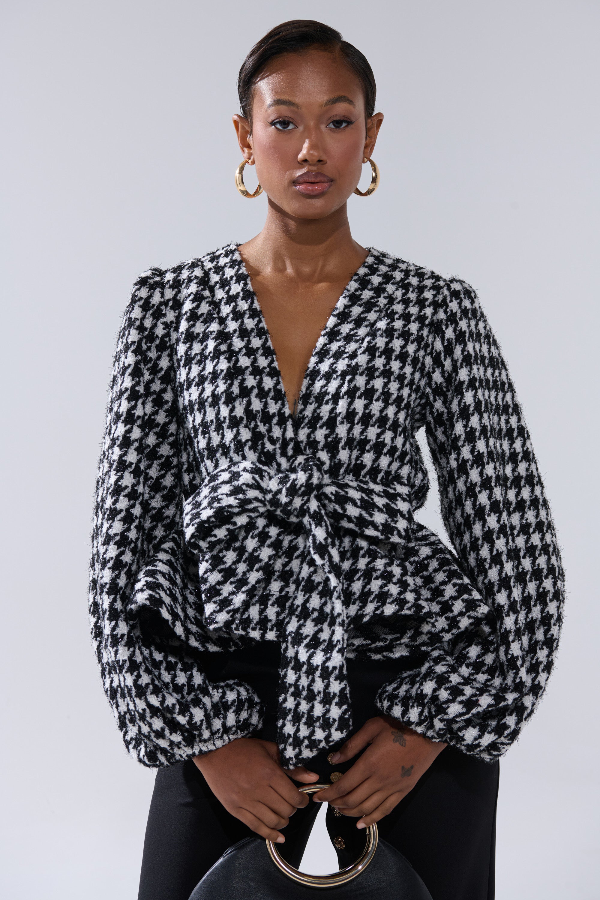 A woman wears the GIRL PLEASE HOUNDSTOOTH TIE-FRONT BLOUSE with a peplum hem, deep V-neck, puffed long sleeves, and waist tie, styled with black pants, hoop earrings, and a black handbag.