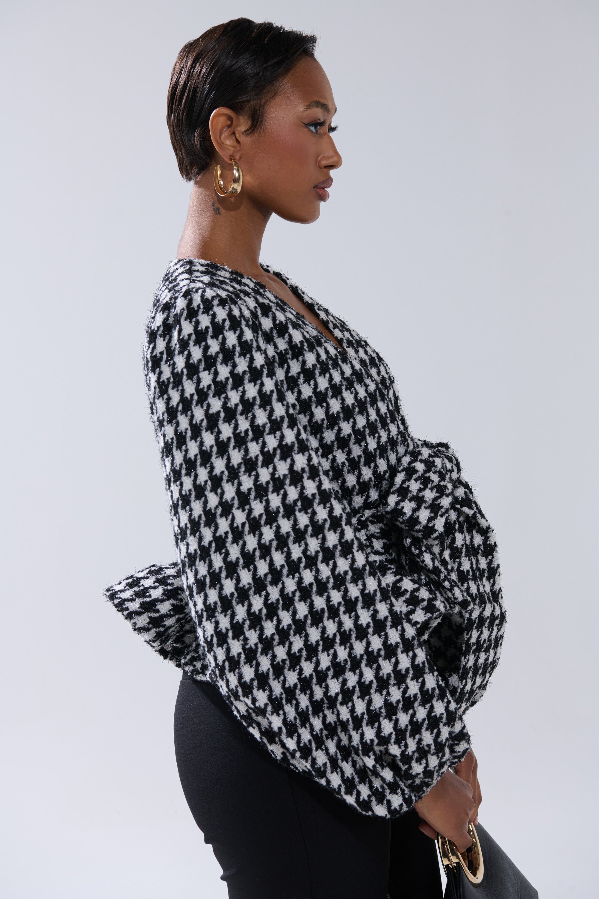 A woman in profile wears the GIRL PLEASE HOUNDSTOOTH TIE-FRONT BLOUSE with voluminous sleeves, large gold hoop earrings, black pants, and holds a black handbag against a plain background.