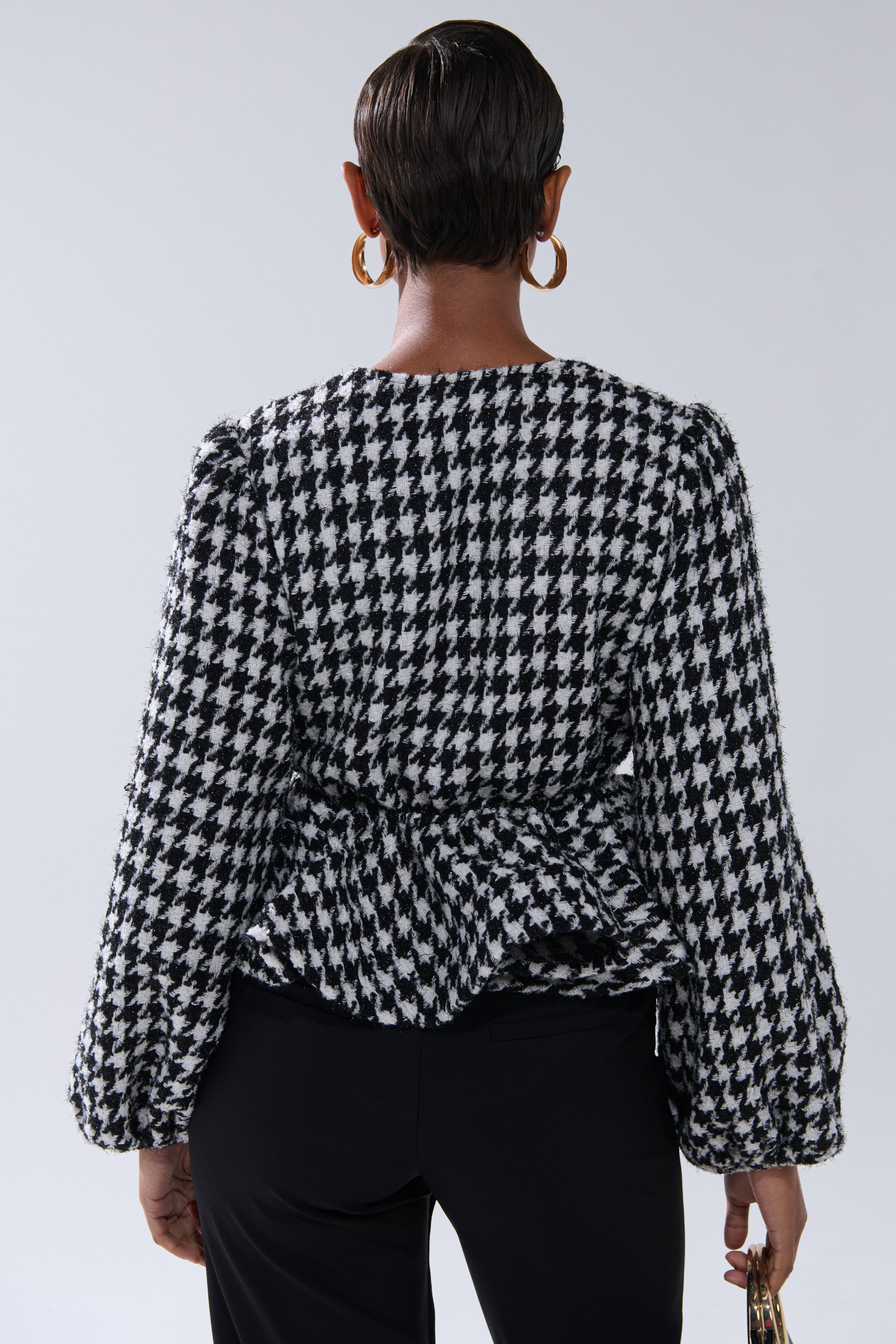 Seen from behind, a person with short hair and large hoop earrings wears the GIRL PLEASE HOUNDSTOOTH TIE-FRONT BLOUSE with puffed sleeves, black pants, and holds a small handbag.