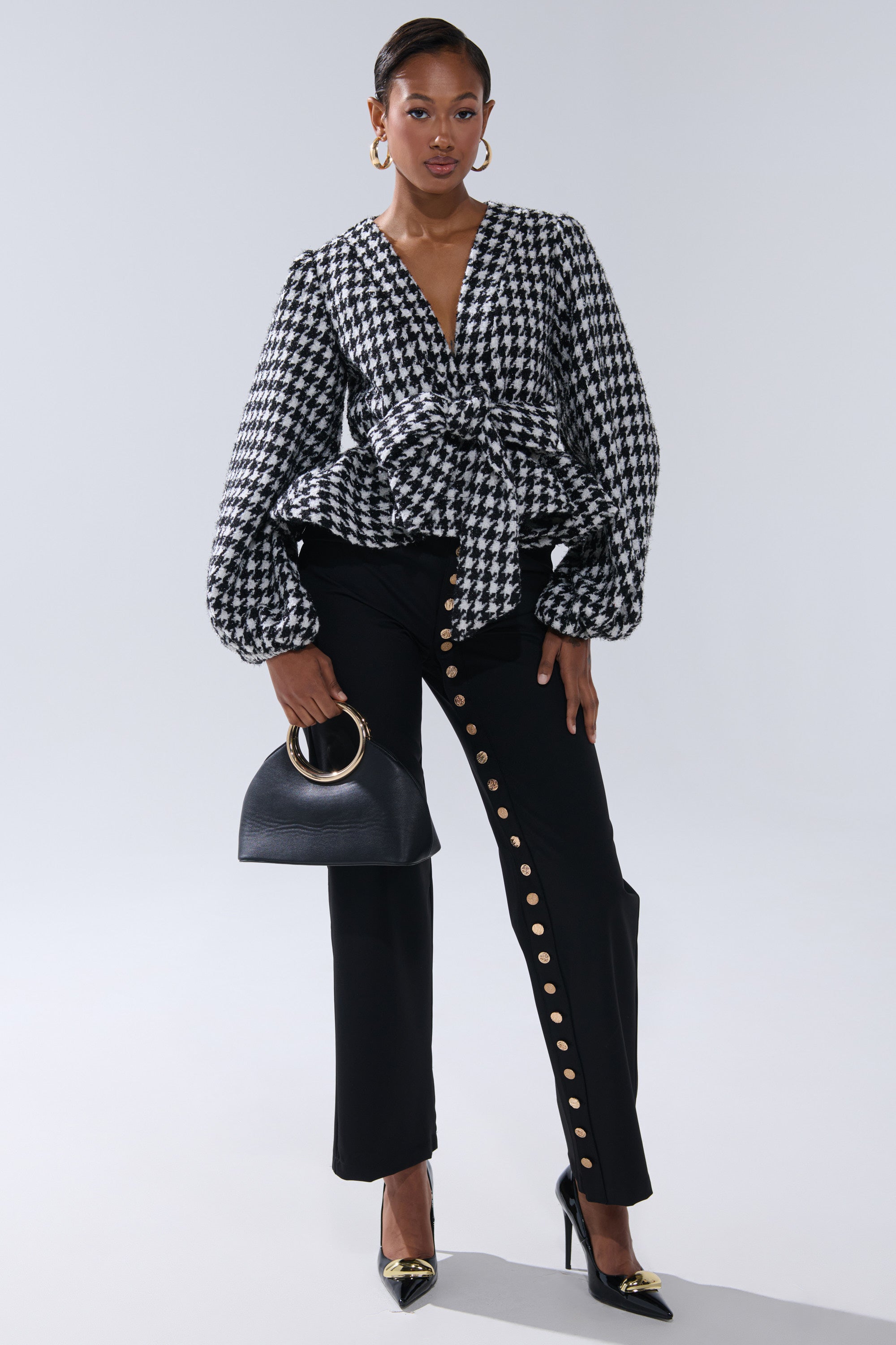 A woman poses confidently in the GIRL PLEASE HOUNDSTOOTH TIE-FRONT BLOUSE, styled with black pants featuring gold side buttons, black heels with gold tips, gold hoop earrings, and a black handbag with a gold handle.