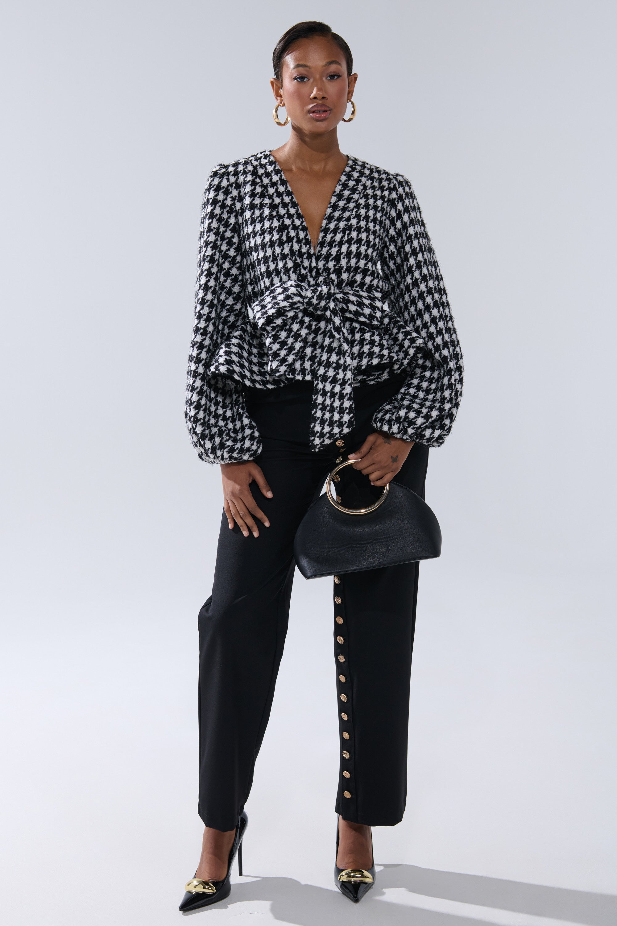 A woman exudes confidence in the GIRL PLEASE HOUNDSTOOTH TIE-FRONT BLOUSE, styled with black pants with gold buttons, black heels with gold tips, and a chic black handbag featuring a gold handle.