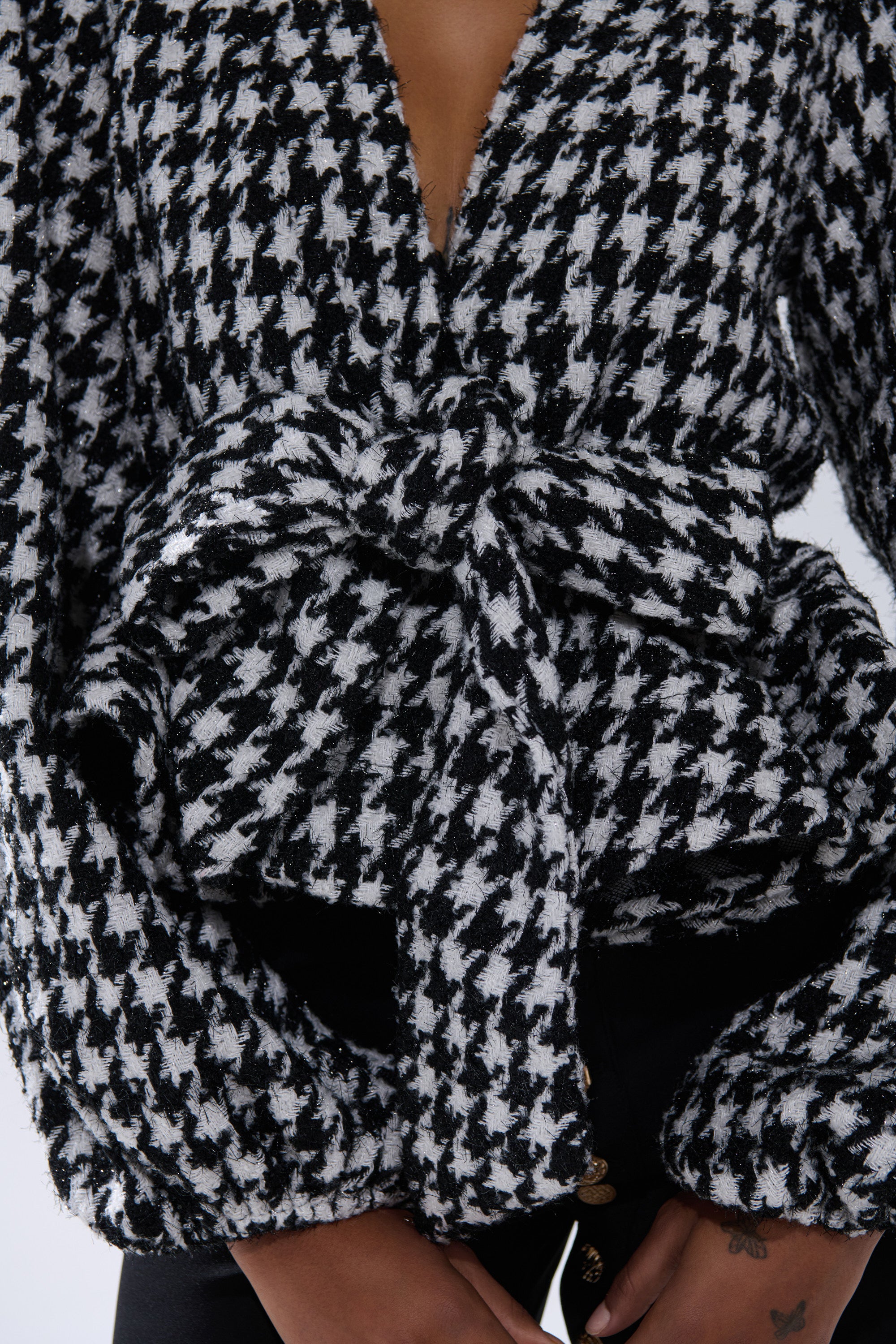 A person wears the GIRL PLEASE HOUNDSTOOTH TIE-FRONT BLOUSE, featuring a black and white houndstooth pattern, deep V-neck, and large waist bow, paired with black pants. Hands appear at the bottom of the image.