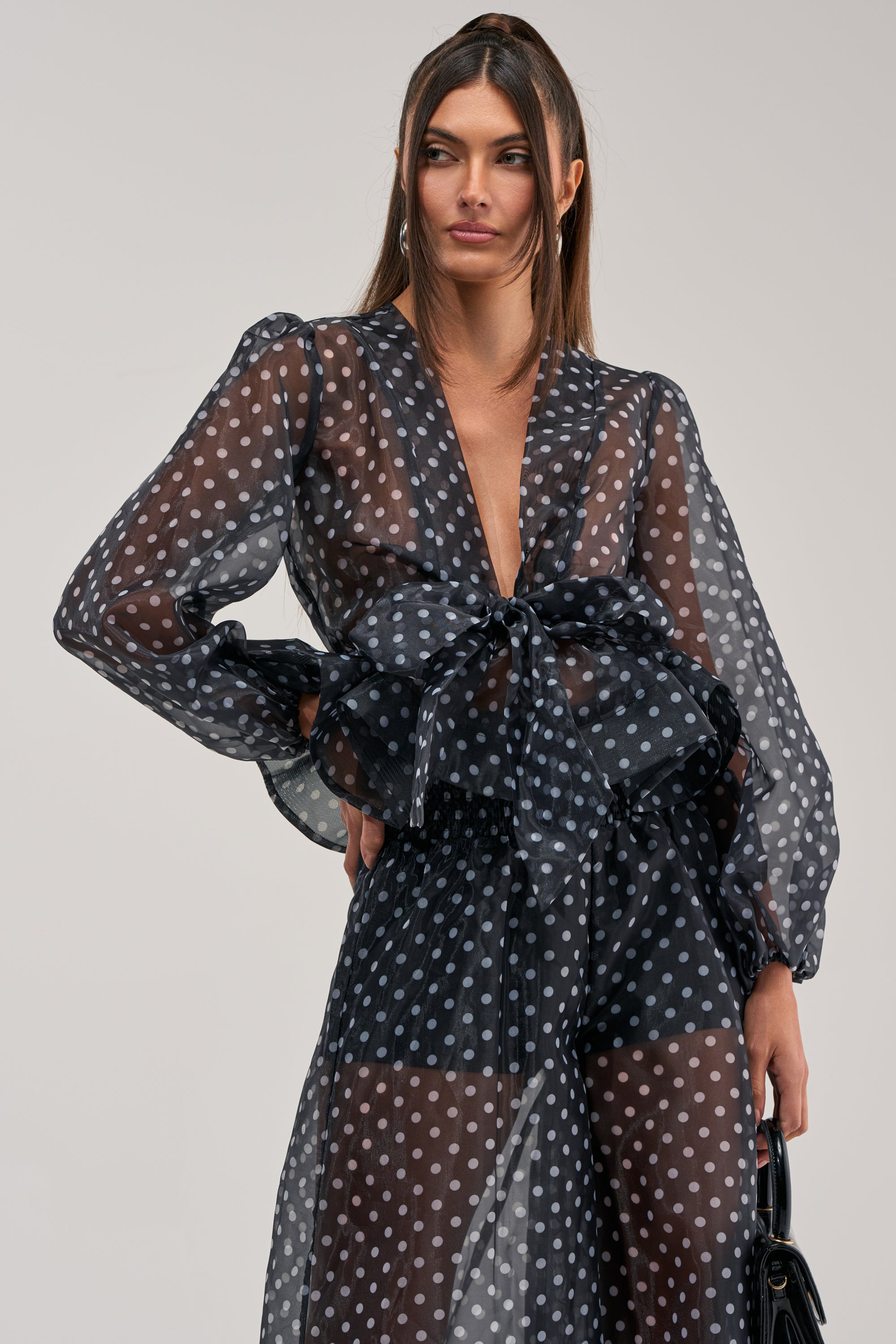 A woman models the PRETTY GIRL POLKA DOT BLOUSE with puffy sleeves and a large waist bow, paired with a sheer skirt. She stands against a light background and holds a black handbag.