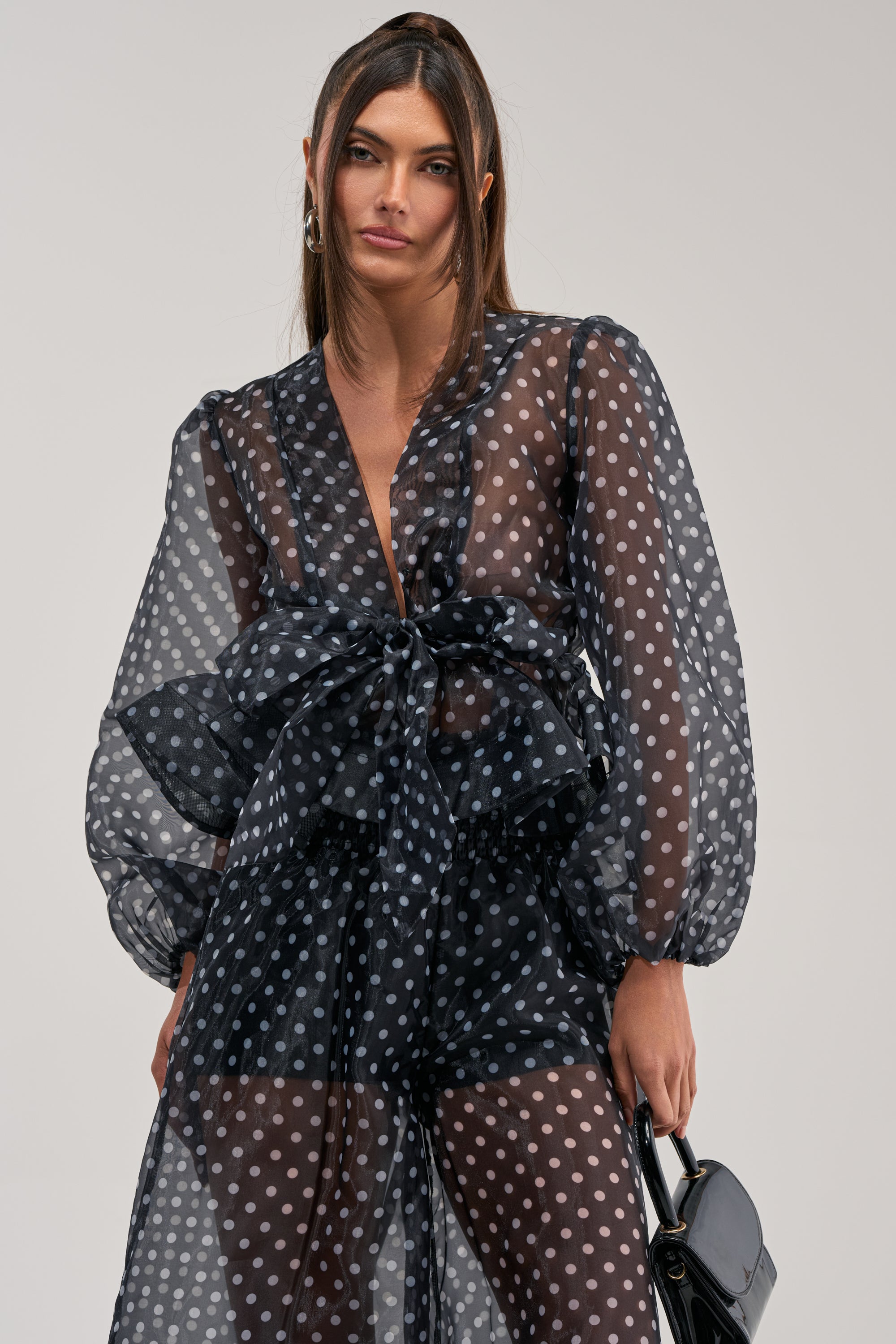 Wearing the PRETTY GIRL POLKA DOT BLOUSE—a sheer black top with white dots, long sleeves, and a large waist bow—a woman poses with a small black handbag, her hair straight and parted in the middle.