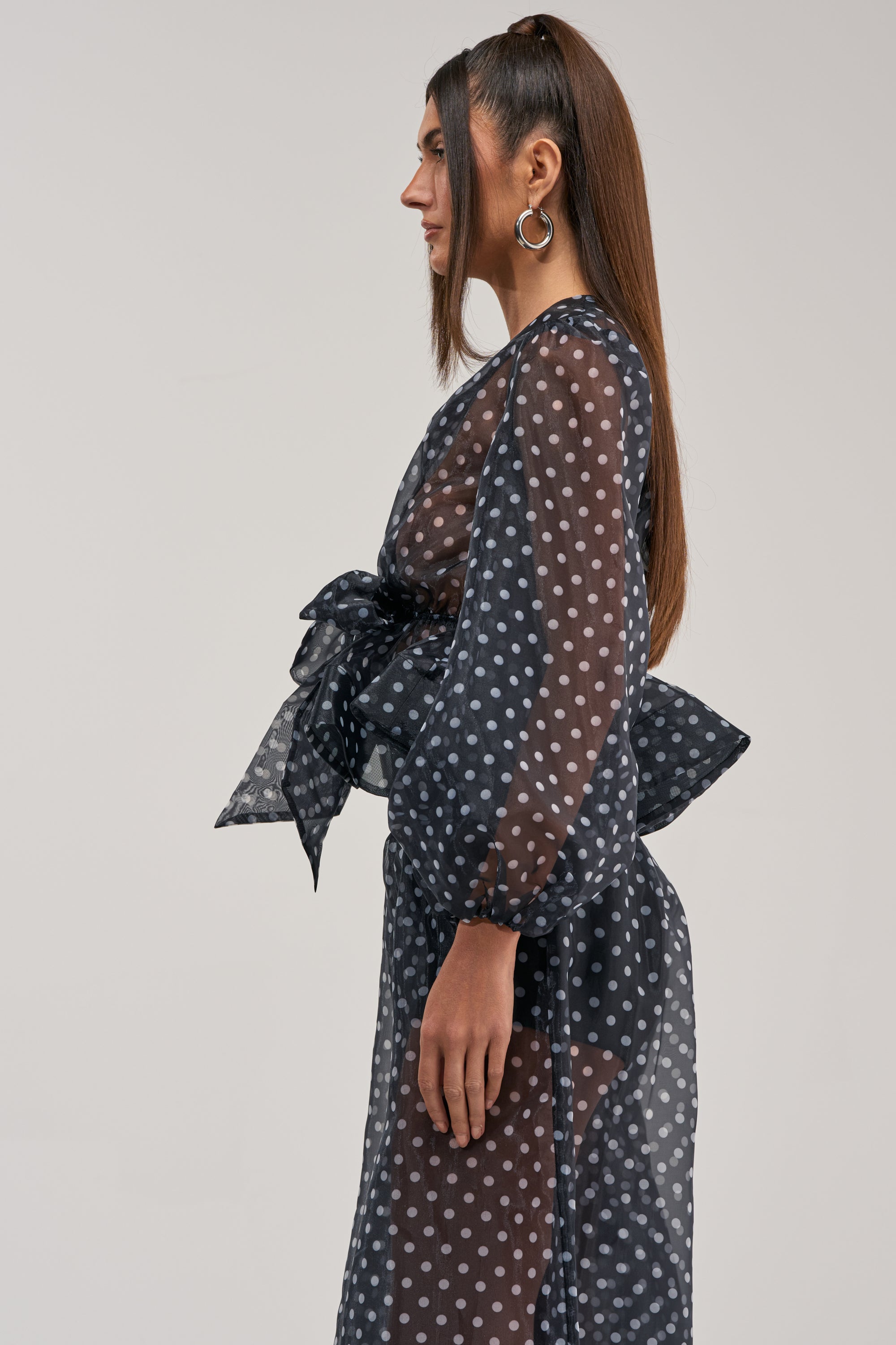 A woman in profile wears the PRETTY GIRL POLKA DOT BLOUSE, a sheer black long-sleeved top styled as a dress with a large waist bow. She has long straight hair, hoop earrings, and stands before a plain light gray background.