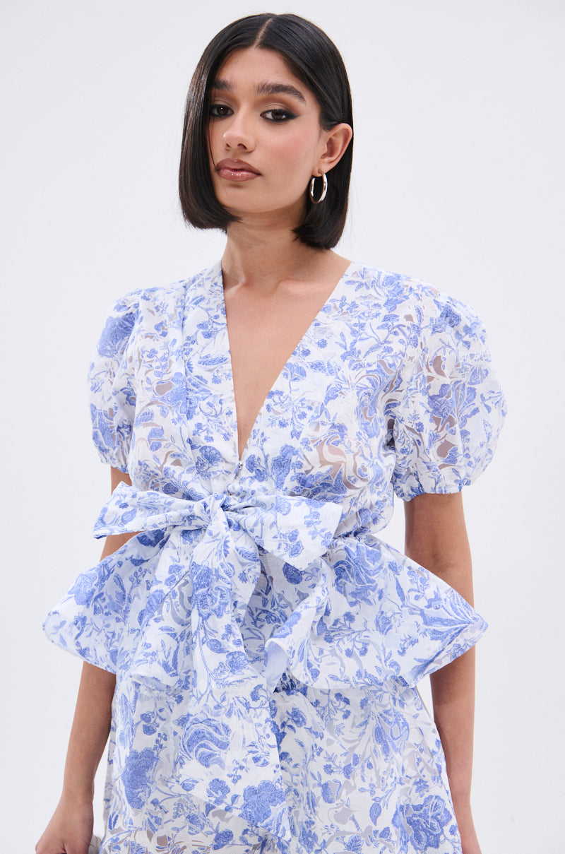 A woman with straight, shoulder-length dark hair wears the BINKEY FLORAL TIE FRONT BLOUSE in light blue and white, featuring a floral pattern, short puff sleeves, deep V-neckline, and a large waist bow, against a plain light background. 0
