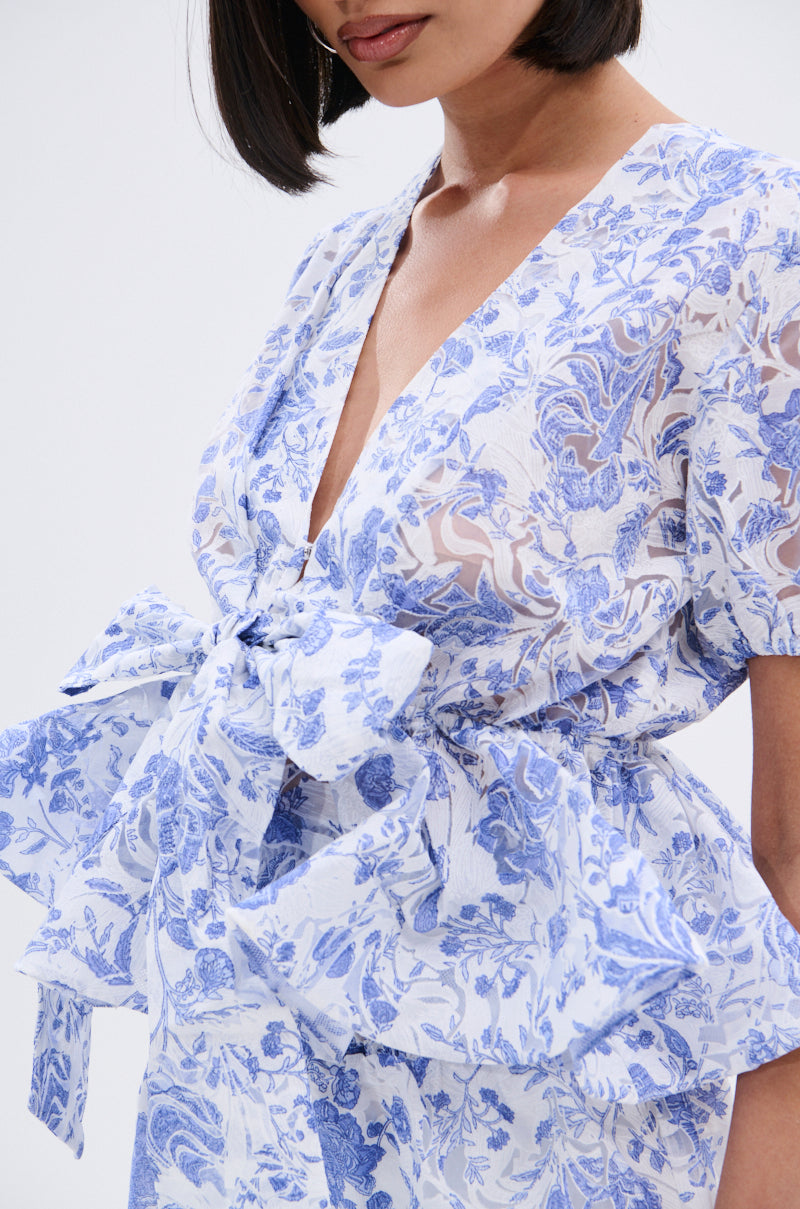 A woman with a short bob wears the BINKEY FLORAL TIE FRONT BLOUSE—white with blue floral patterns, a large bow at the waist, deep V-neckline, and short sleeves. Only her upper body is shown against a plain background. 9