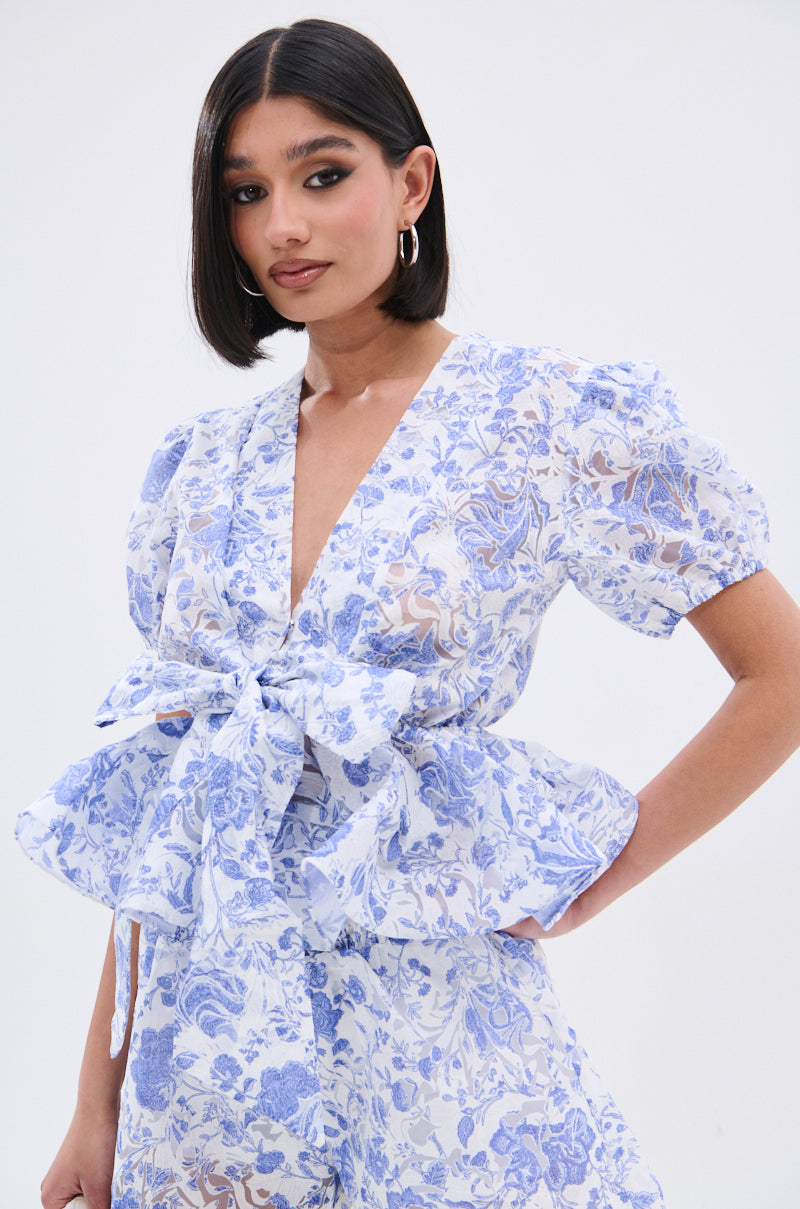 A woman with straight dark hair wears the BINKEY FLORAL TIE FRONT BLOUSE, featuring a light blue and white floral print, puffed short sleeves, a deep V-neckline, and a large tie-front bow. She poses confidently against a plain white background. 1