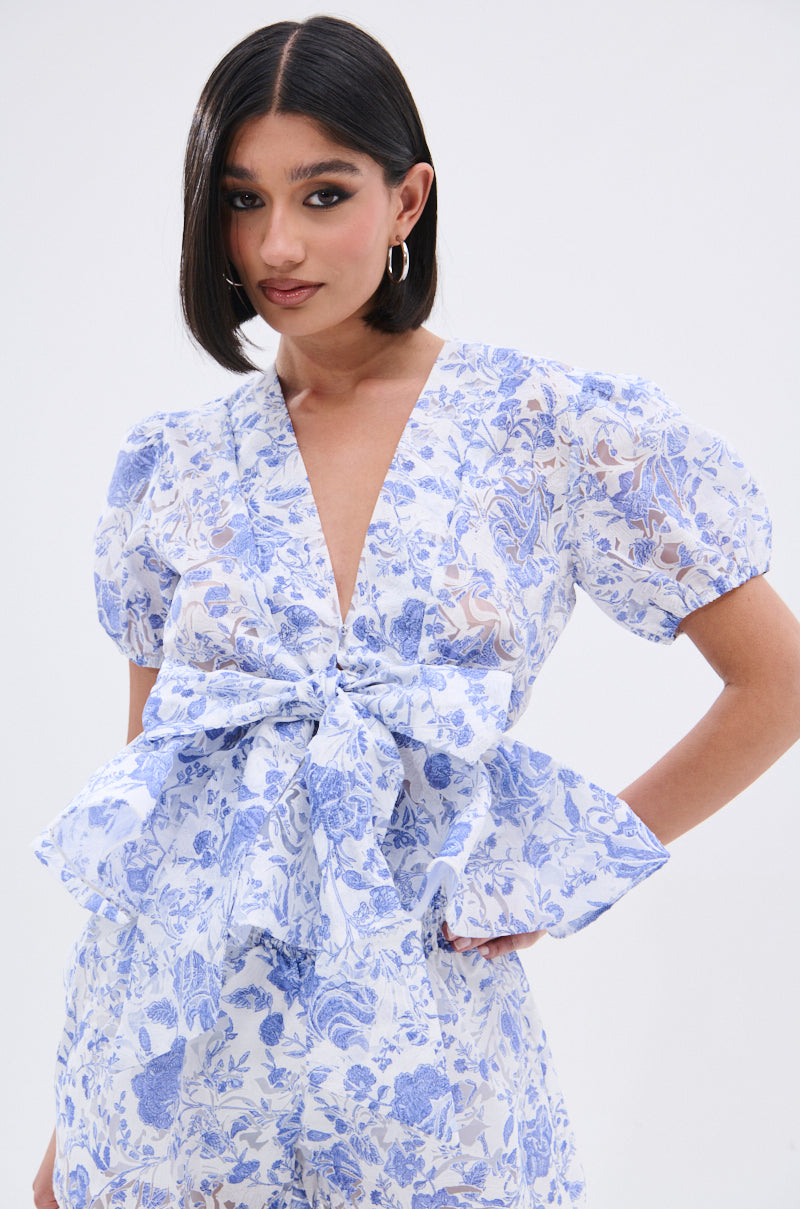A woman with a sleek bob wears the BINKEY FLORAL TIE FRONT BLOUSE, a light blue and white floral top featuring puffed short sleeves, a deep V-neckline, and a tie-front bow, standing against a plain light background. 2