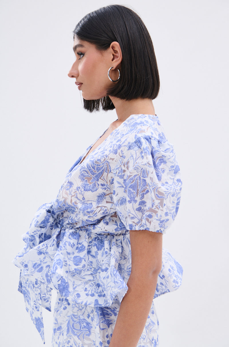 A woman with a sleek bob showcases the BINKEY FLORAL TIE FRONT BLOUSE, featuring blue and white floral print, puffed short sleeves, and a large waist bow, as she stands in profile against a plain light background. 3