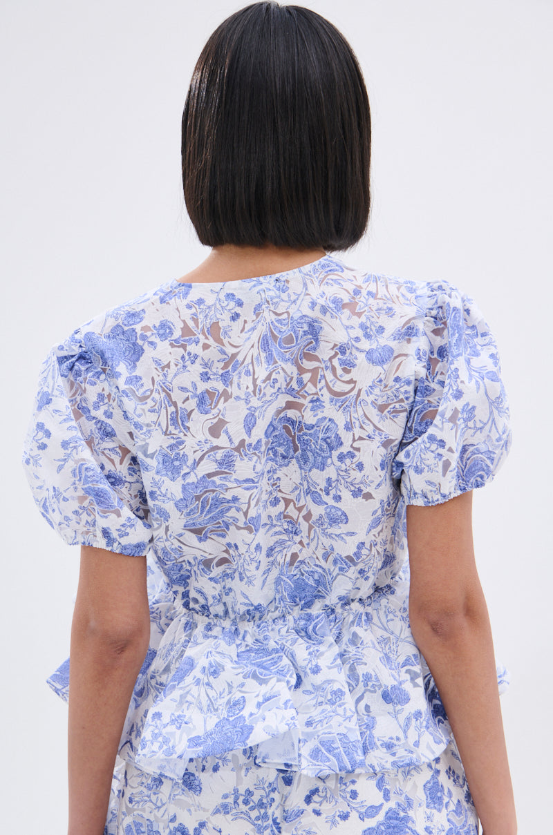 A person with straight, shoulder-length dark hair is seen from behind wearing the BINKEY FLORAL TIE FRONT BLOUSE, featuring a blue and white floral pattern, sheer lace on the upper back, and delicate ruffles. 4