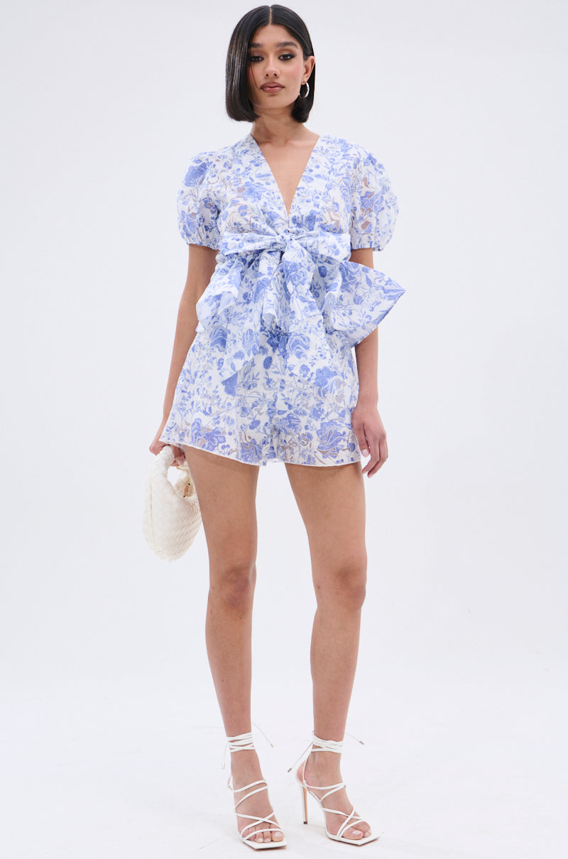 A person wears the BINKEY FLORAL TIE FRONT BLOUSE in light blue and white with puff sleeves and a peplum silhouette, standing against a plain background, holding a white textured clutch and wearing strappy white heels. 7