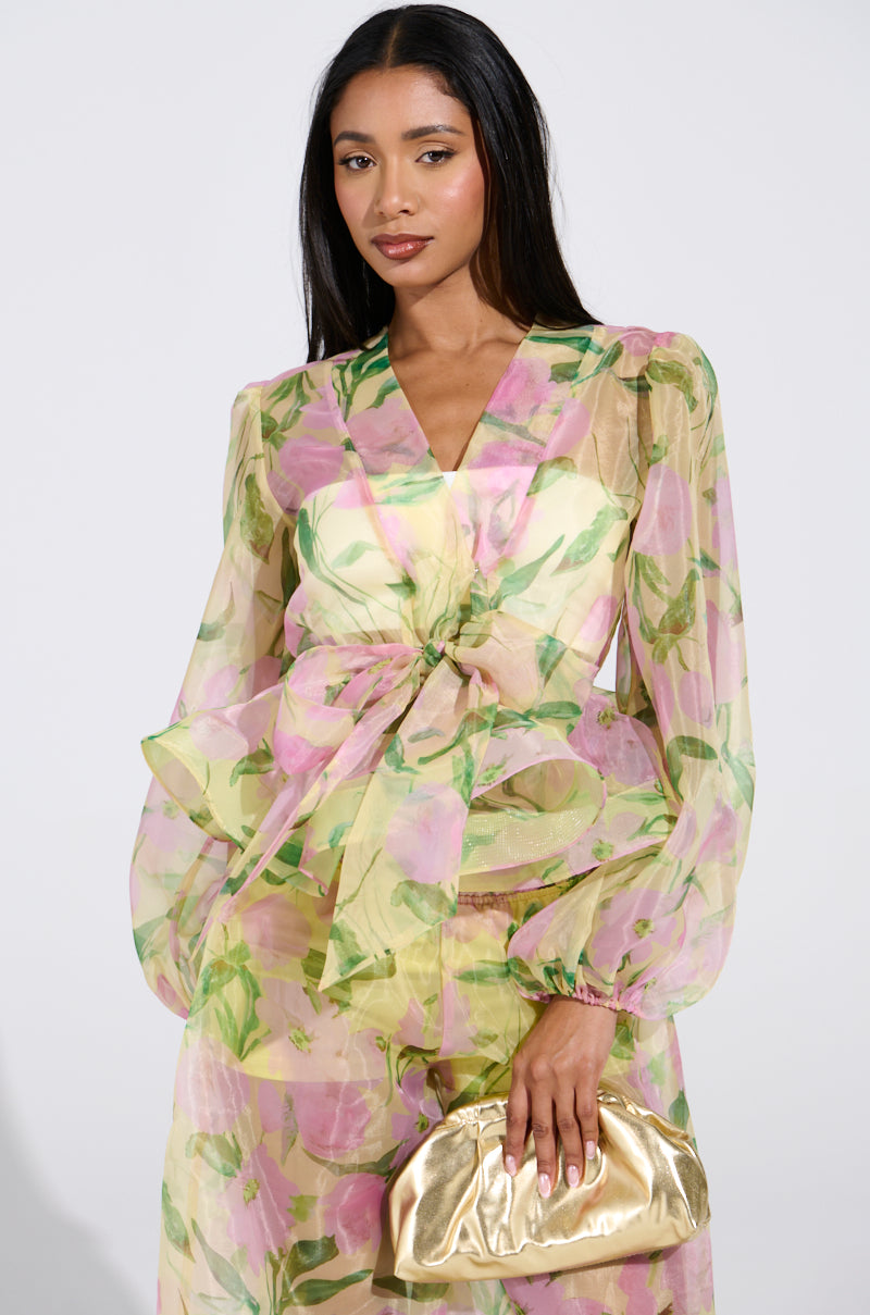 A woman wearing the FLOWER POWER CHIFFON TIE-FRONT BLOUSE, adorned with pink and green botanical floral prints, holds a gold clutch. The blouse's elegant design features long sheer sleeves and a bow at the waist. Her long dark hair cascades against a plain light background.