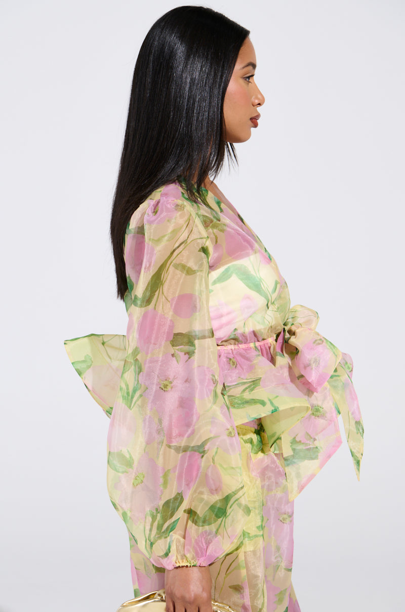 A person with long black hair wears a flowing, semi-transparent floral outfit showcasing a Botanical Floral Print in shades of pink and green. Resembling the FLOWER POWER CHIFFON TIE-FRONT BLOUSE, it features a large bow at the waist. A gold purse is held as they face sideways.