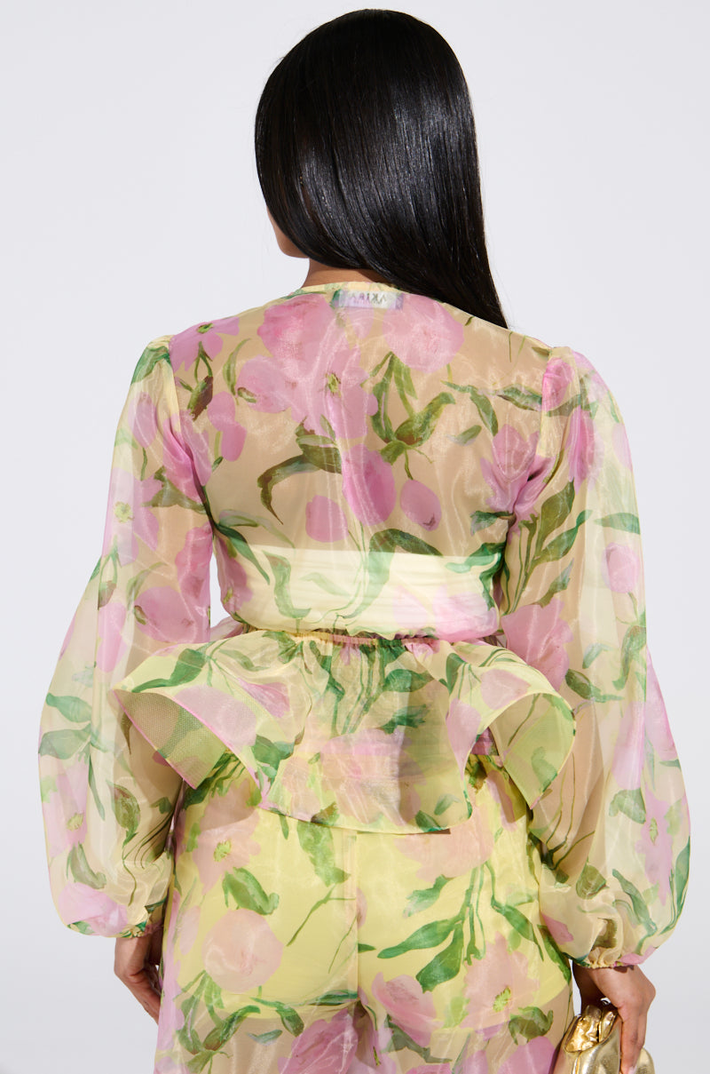 A woman with long black hair wears a FLOWER POWER CHIFFON TIE-FRONT BLOUSE, showcasing pink and green botanical floral prints, puffed sleeves, and a peplum waist. She holds a small gold purse, facing away against a plain backdrop.