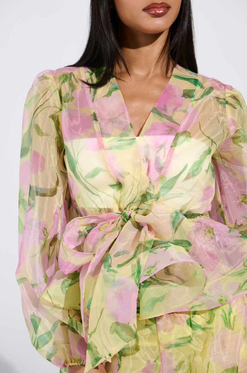 A woman wears the FLOWER POWER CHIFFON TIE FRONT BLOUSE, featuring a floral print with sheer organza and long sleeves. The pink and green blend is enhanced by a waist bow. Her straight dark hair cascades over her shoulders against a plain white background.