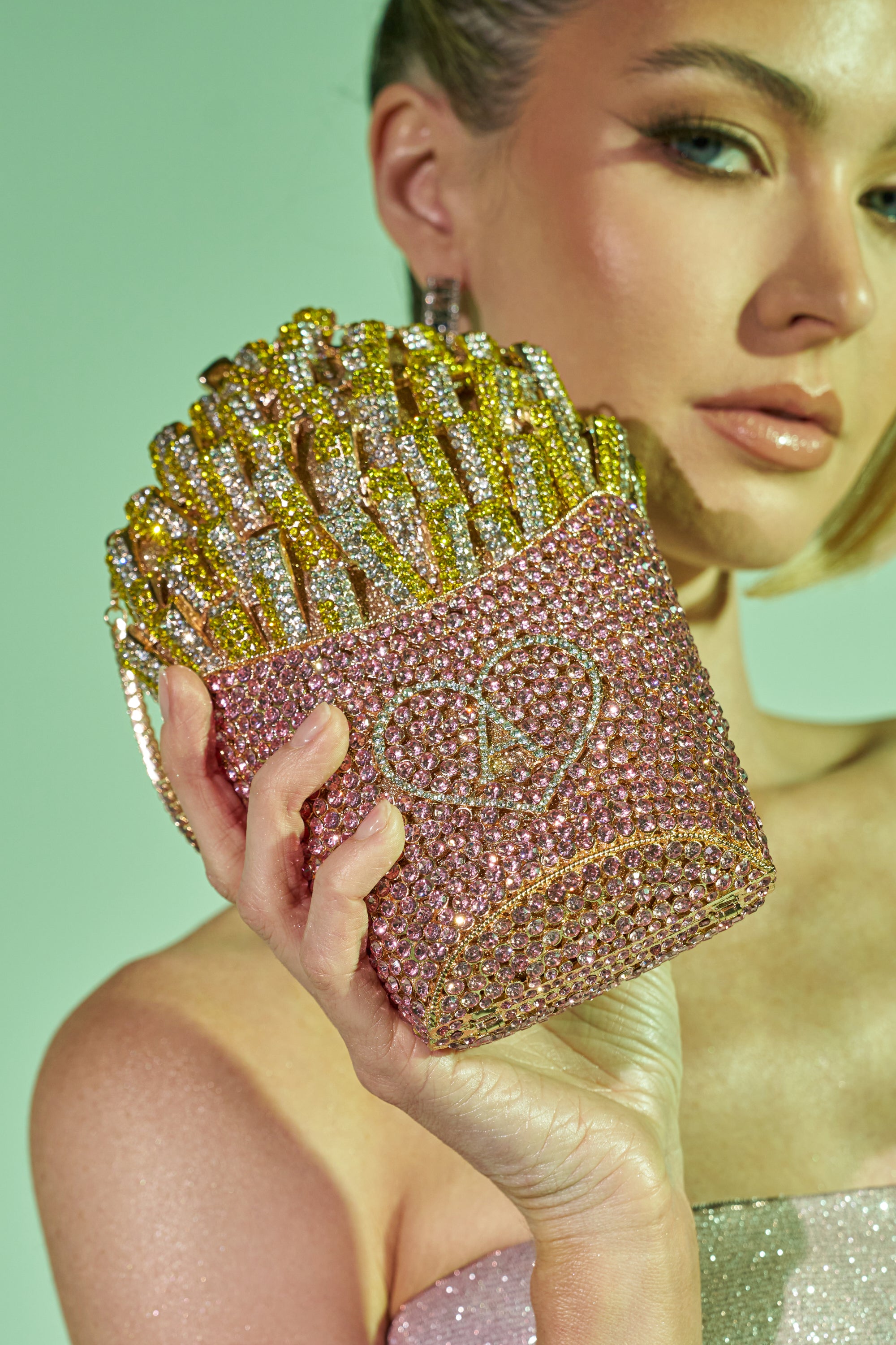 A woman holds the A-MAZING FRENCH FRY BLING PURSE, a crystal-embellished clutch shaped like a box of fries with gold and silver accents. She wears shimmering makeup and a glittery strapless top against a vibrant green background. 0