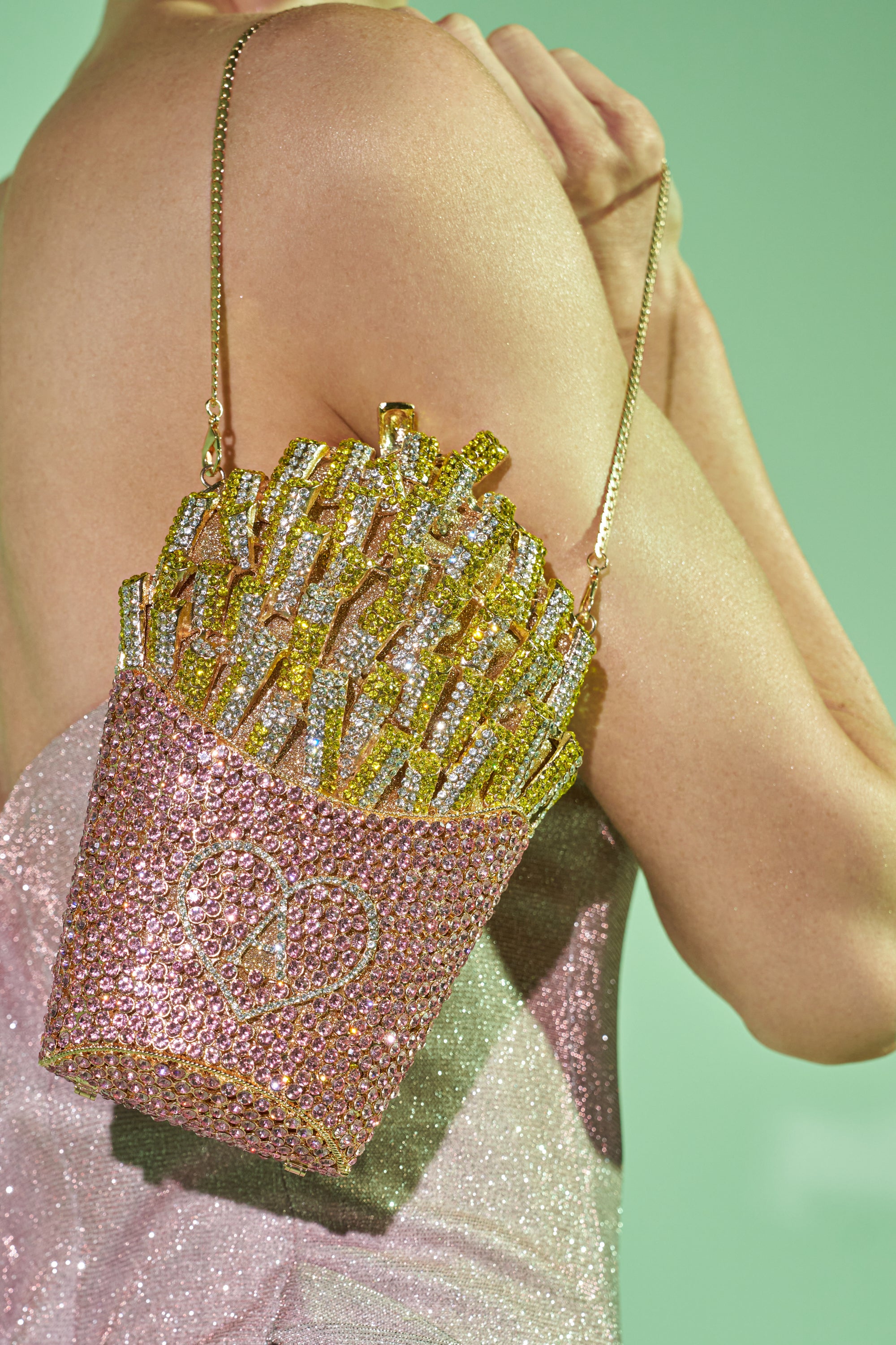 A person in a sparkly light pink dress holds the A-MAZING FRENCH FRY BLING PURSE—a novelty bag covered in pink, gold, and silver crystals—against a light green background. 1