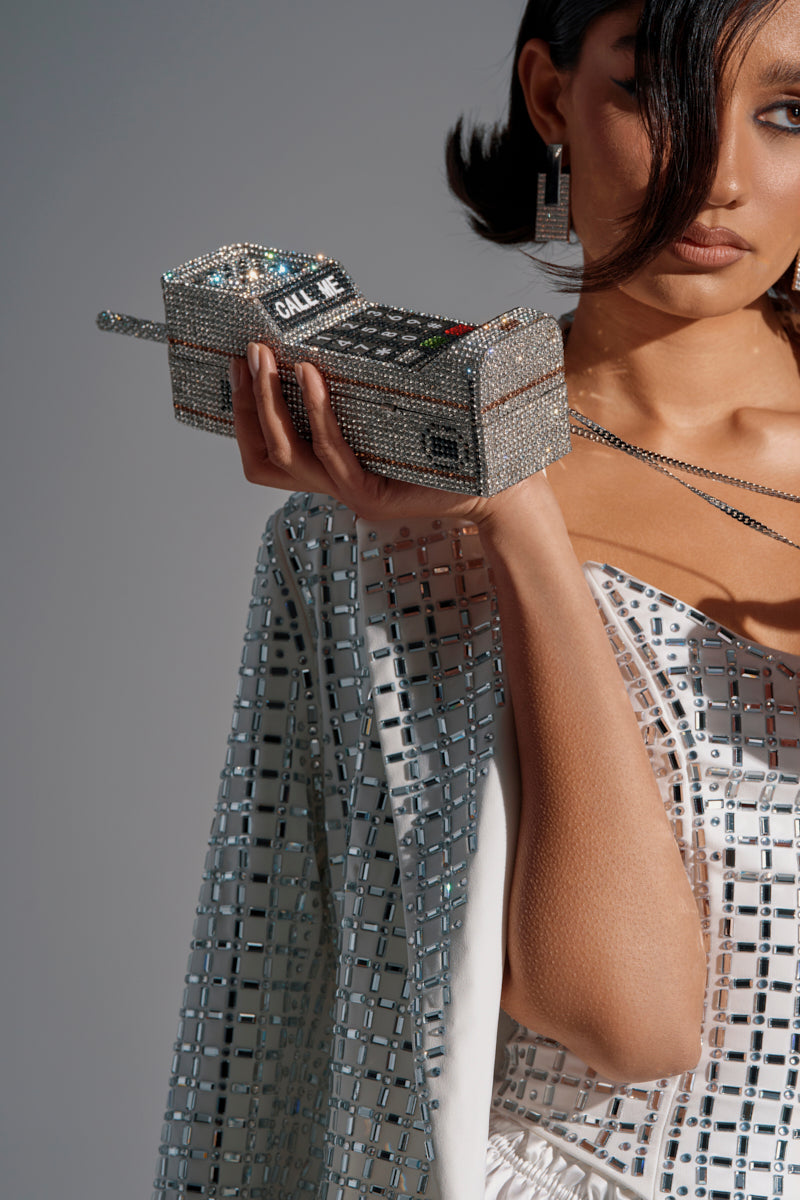 A woman with dark hair and geometric earrings holds the CALL ME BLING CLUTCH, a sparkling, rhinestone phone-shaped handbag, while wearing a silver geometric-patterned outfit. Her face is partially visible.