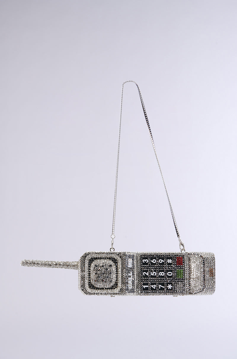 Introducing the CALL ME BLING CLUTCH, a phone-shaped purse designed to resemble a vintage mobile phone with an antenna and numeric keypad. Embellished with sparkling rhinestones and featuring a silver chain strap, this stylish accessory stands out against its plain gray background.